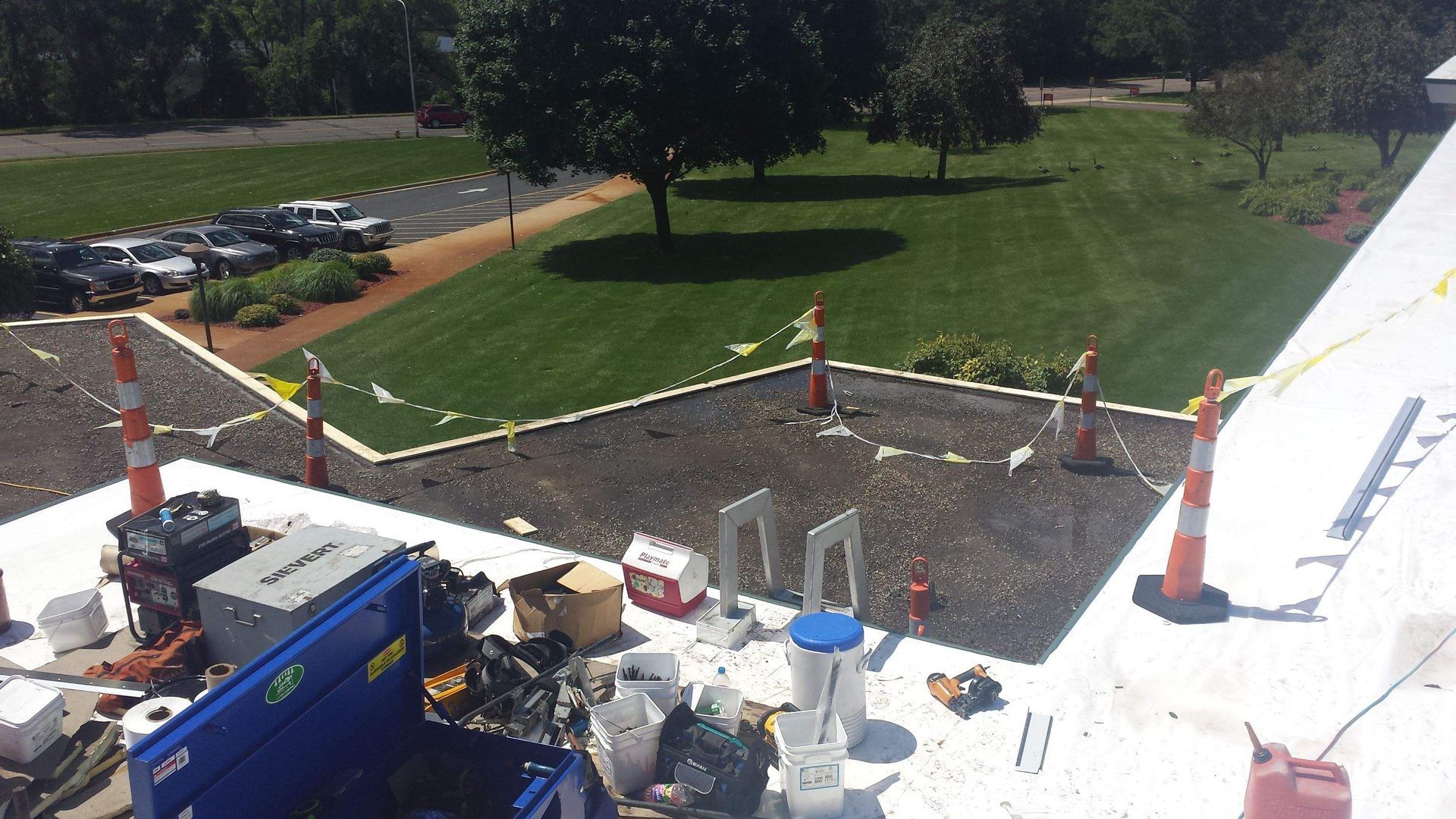 A view from a rooftop looking down at construction materials and equipment, with a lawn and parking lot in the background.