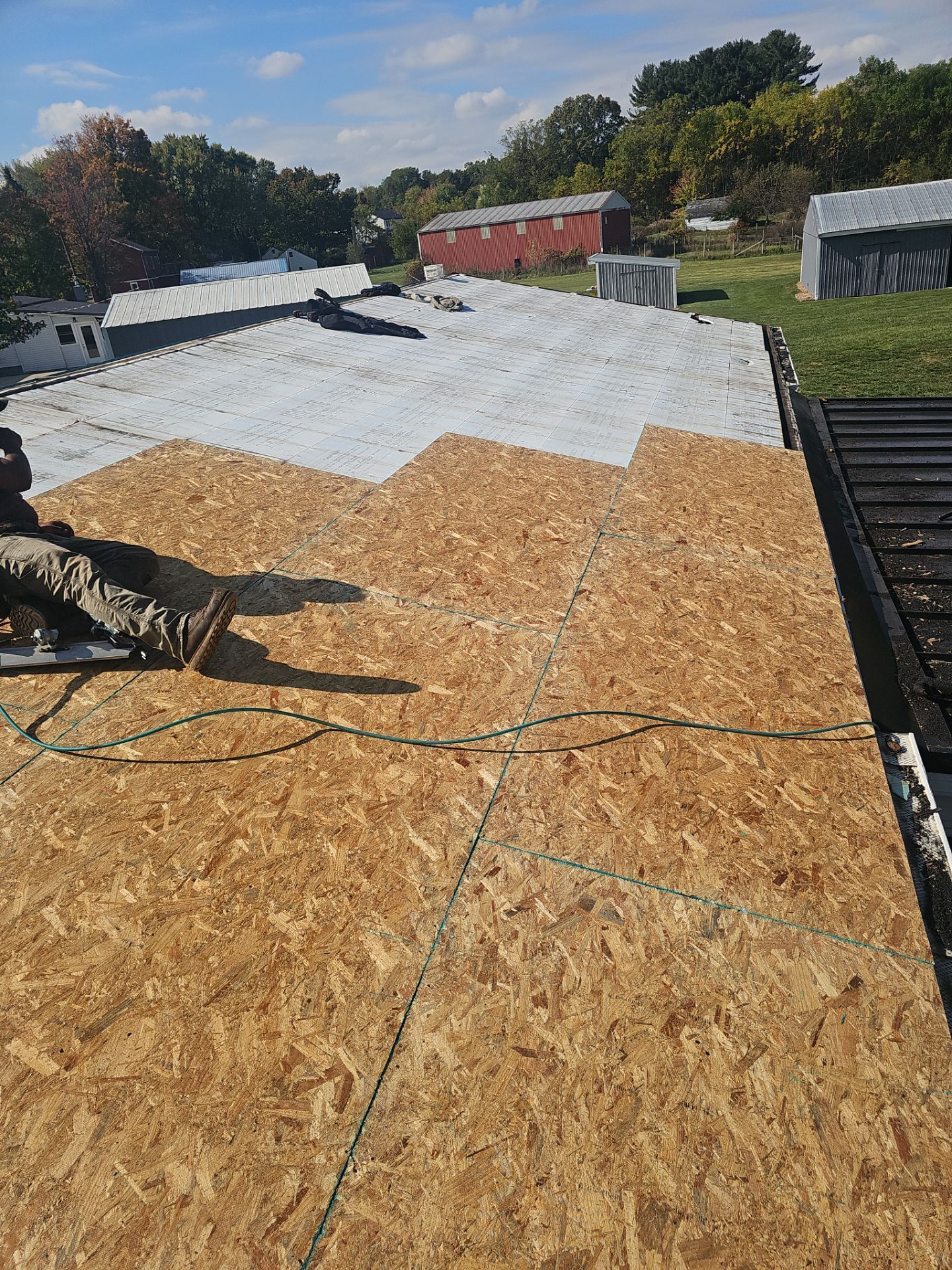 A worker kneeling on a partially shingled roof, installing new wooden plywood decking.