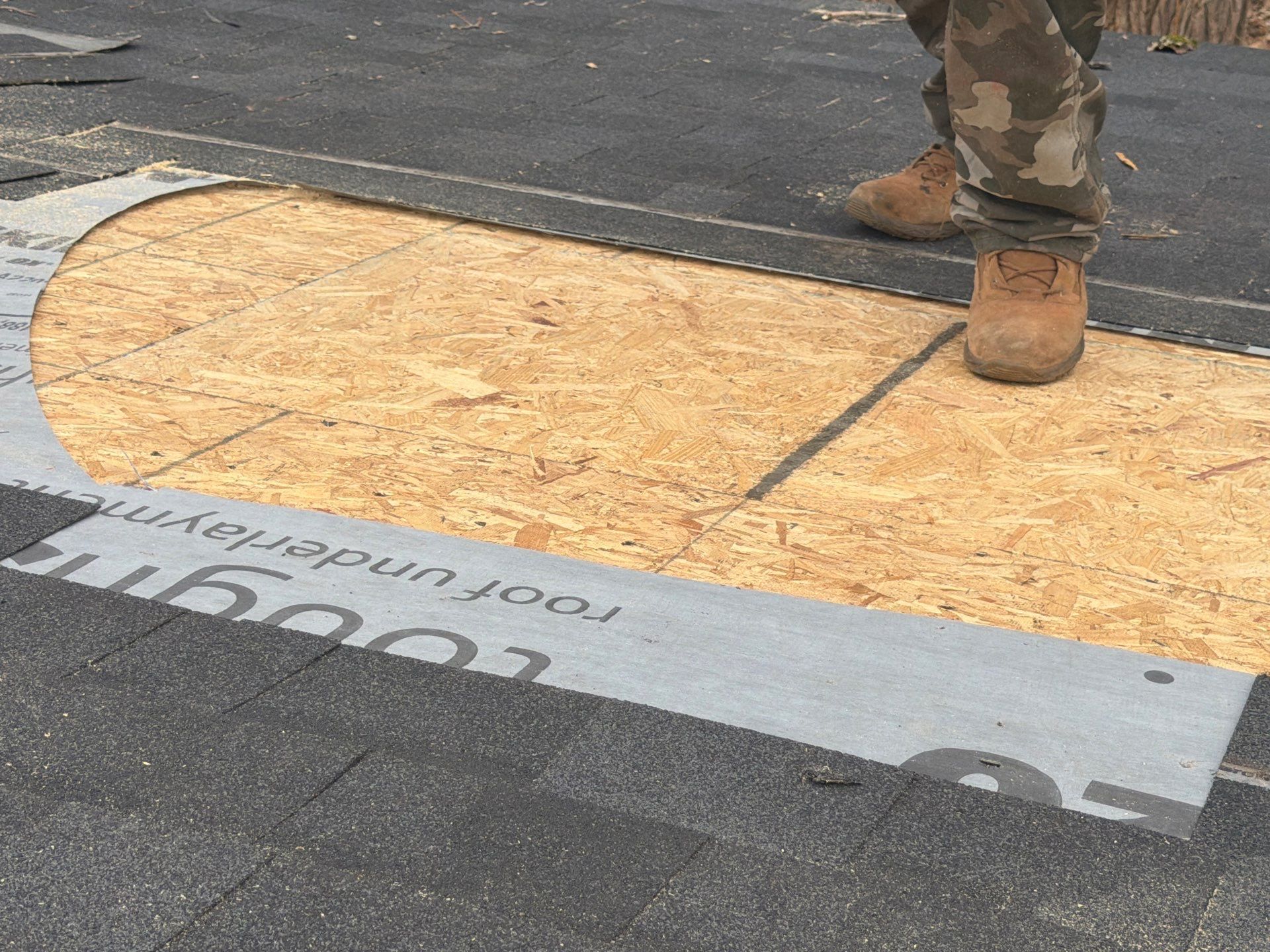 A construction worker stands on a roof deck where a circular section of plywood sheathing is exposed amidst dark shingles.