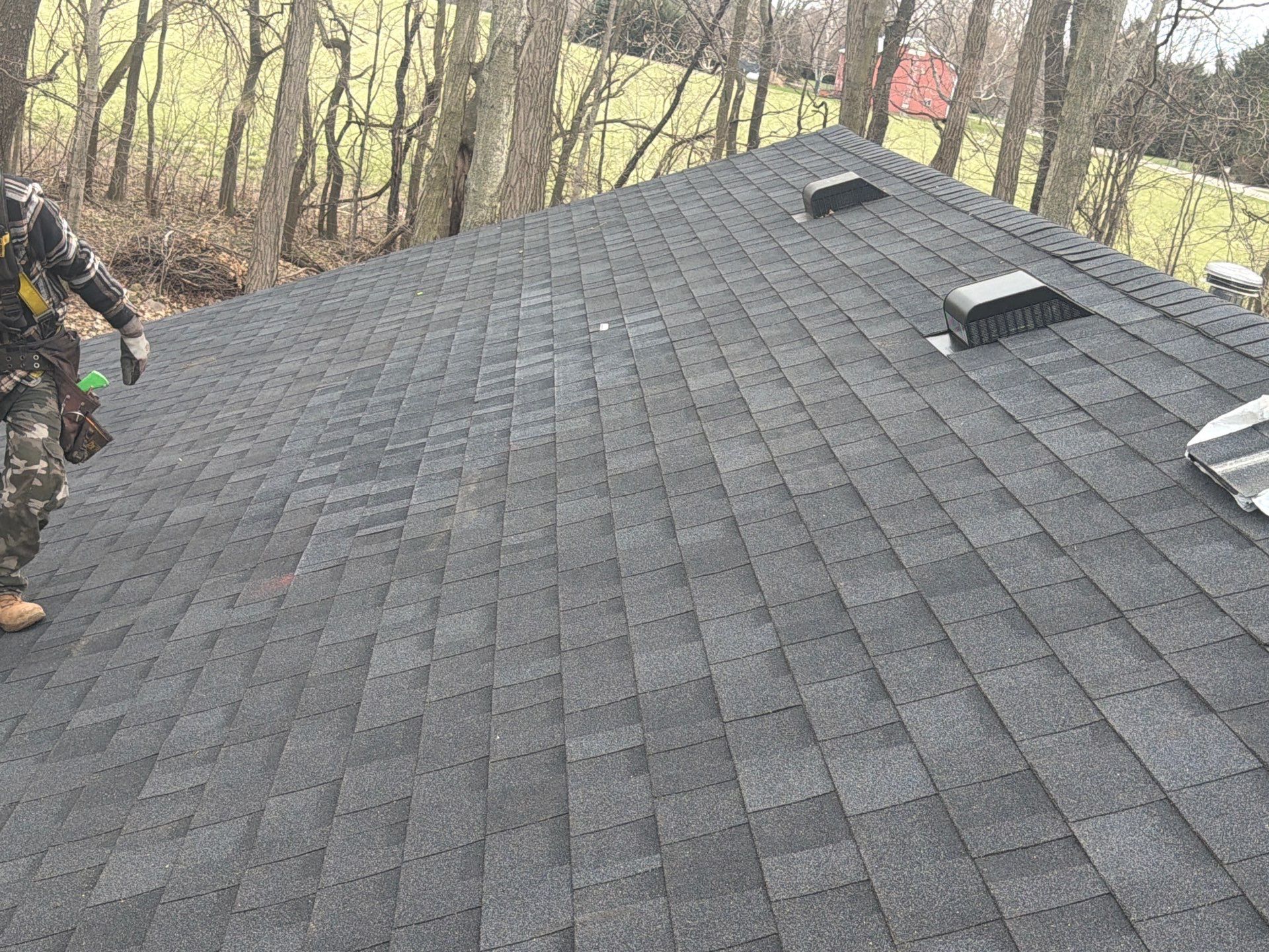 A roofer wearing a safety harness stands on a residential roof with gray asphalt shingles and two black ventilation vents.