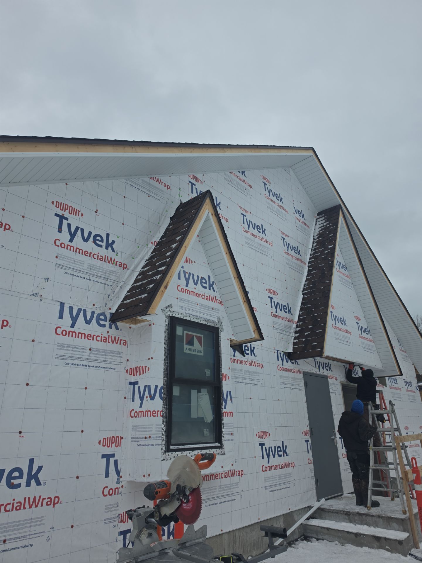 Two workers on a ladder installing building materials on the exterior of a house covered in white Tyvek wrap.