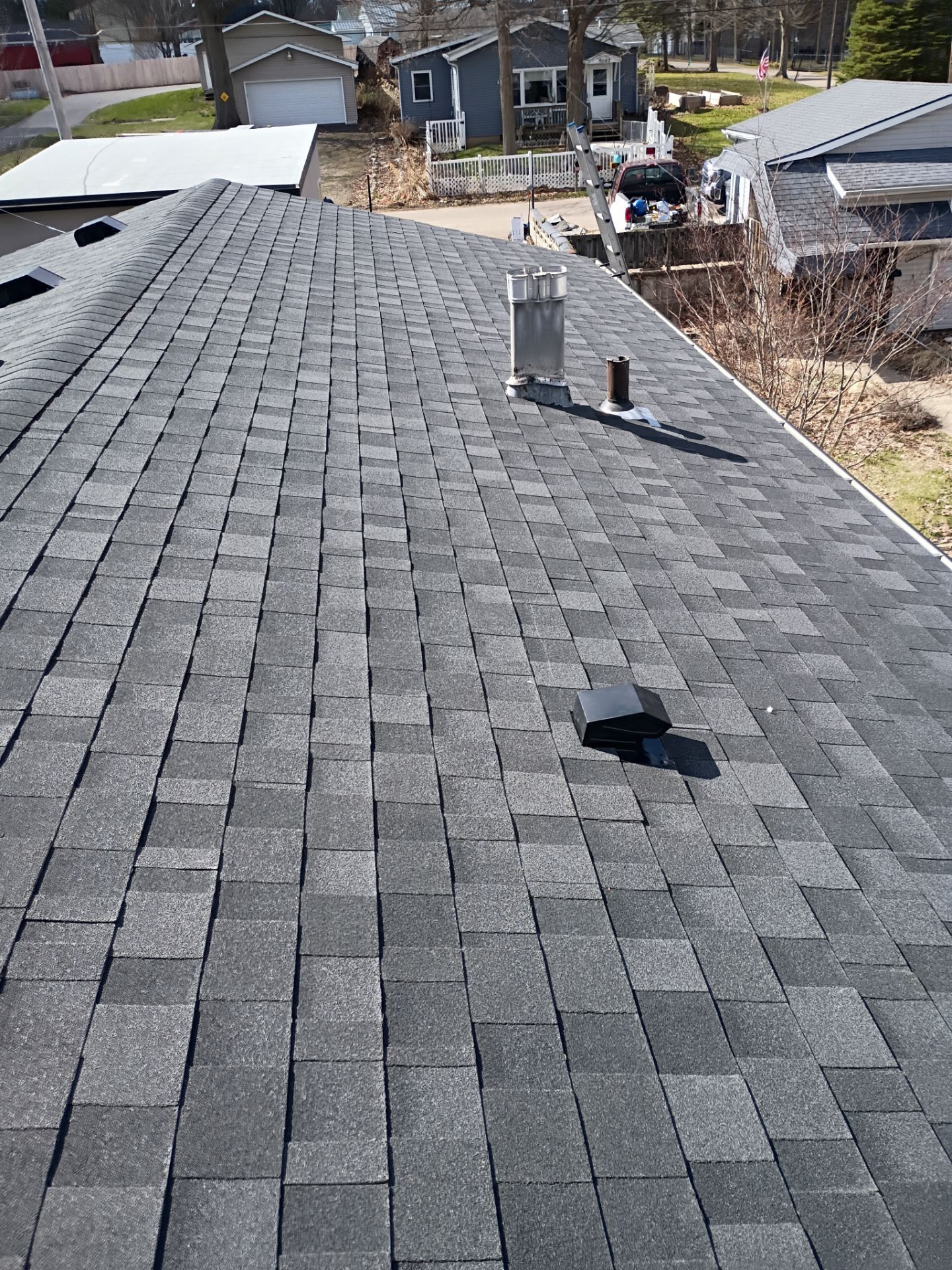 A high-angle view of a grey shingled roof with a chimney, a plumbing vent, and a black exhaust vent.