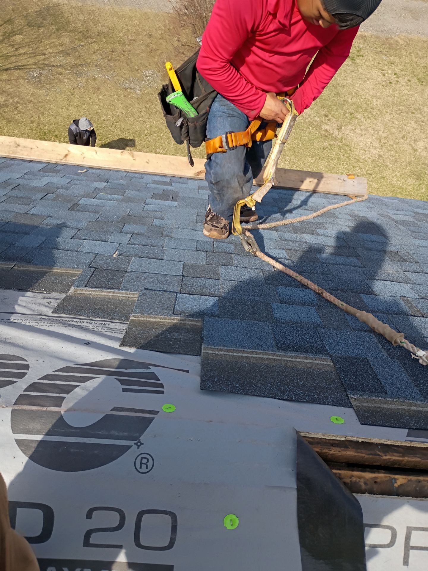 A person in a red shirt wears a safety harness and secures a lifeline while working on a partially shingled roof.