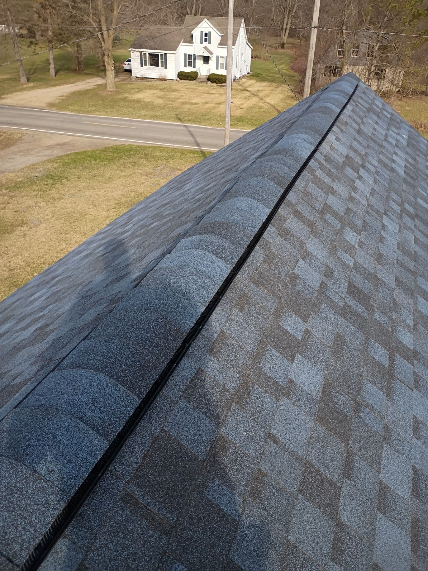 A high-angle view of a gray asphalt shingle roof, focusing on the ridge cap shingles, with a house visible in the distance.