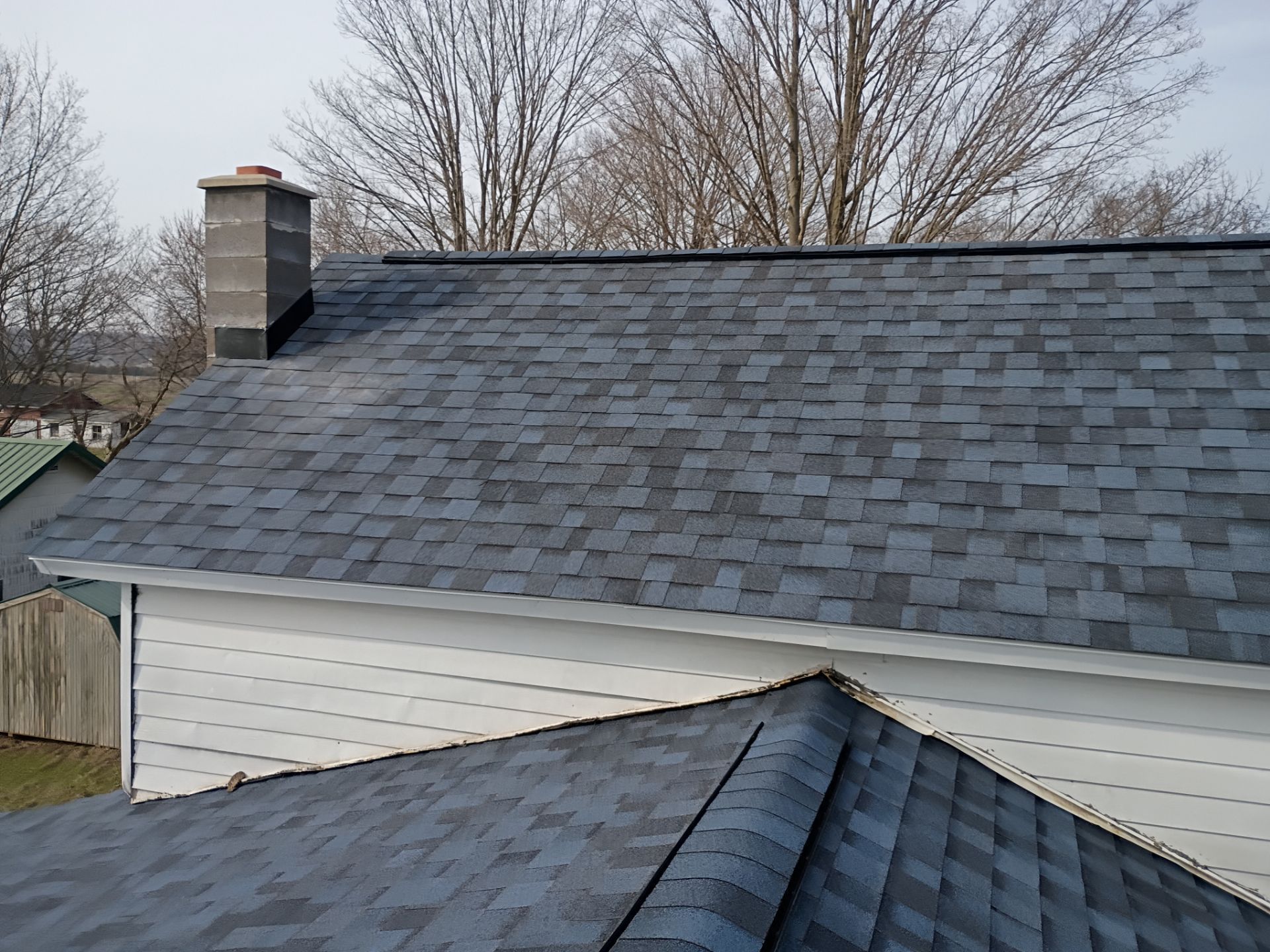 A view of a house roof featuring blue-gray architectural shingles, a chimney, and white siding under a clear sky.