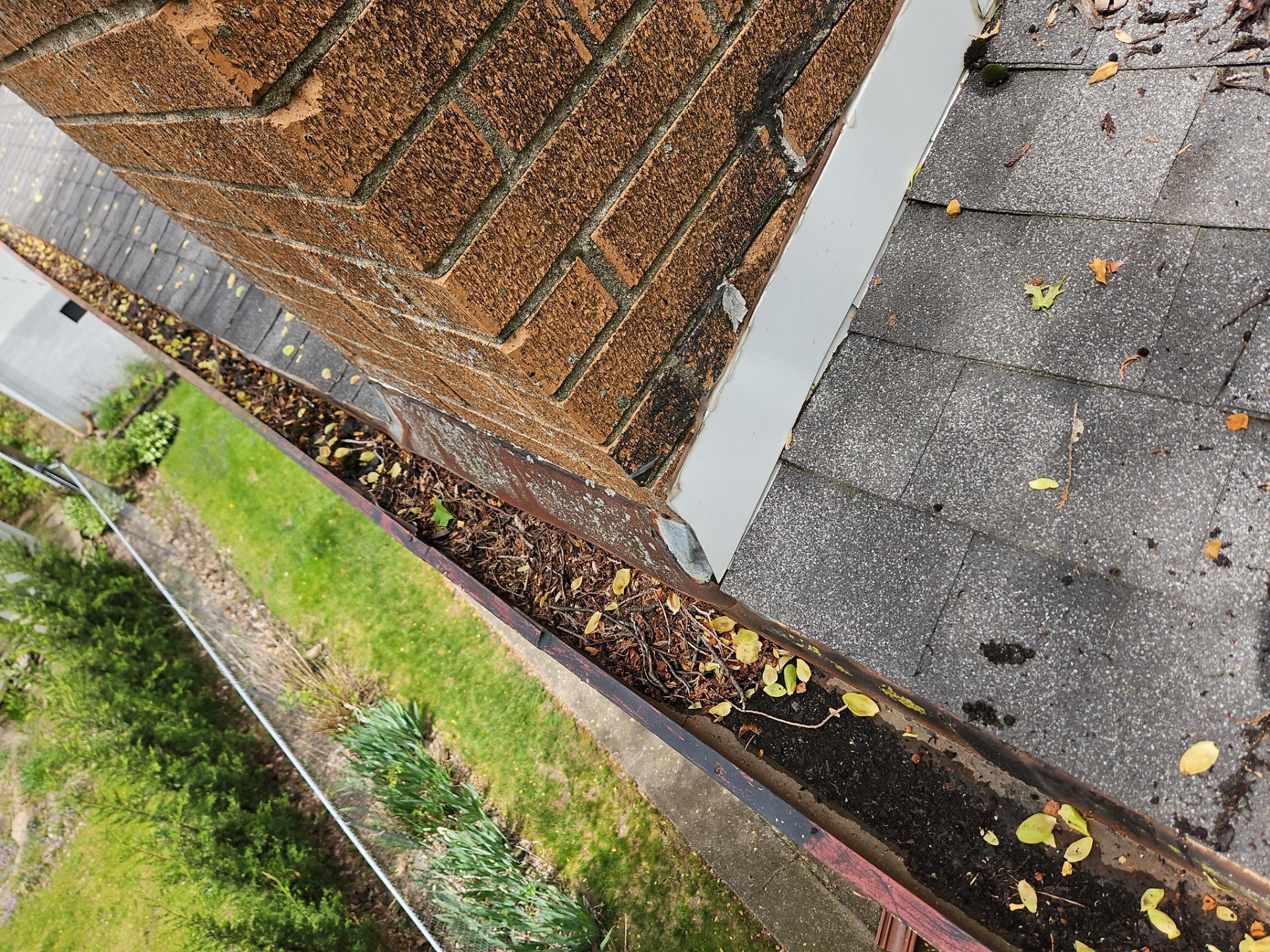 A high-angle view of a brown brick chimney and grey roof shingles, showing a gutter filled with debris and dark dirt.