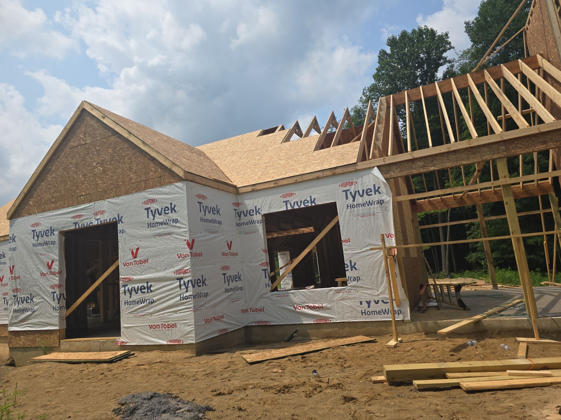 Construction of a house showing wooden framing, a plywood roof, and white Tyvek house wrap against a cloudy sky.