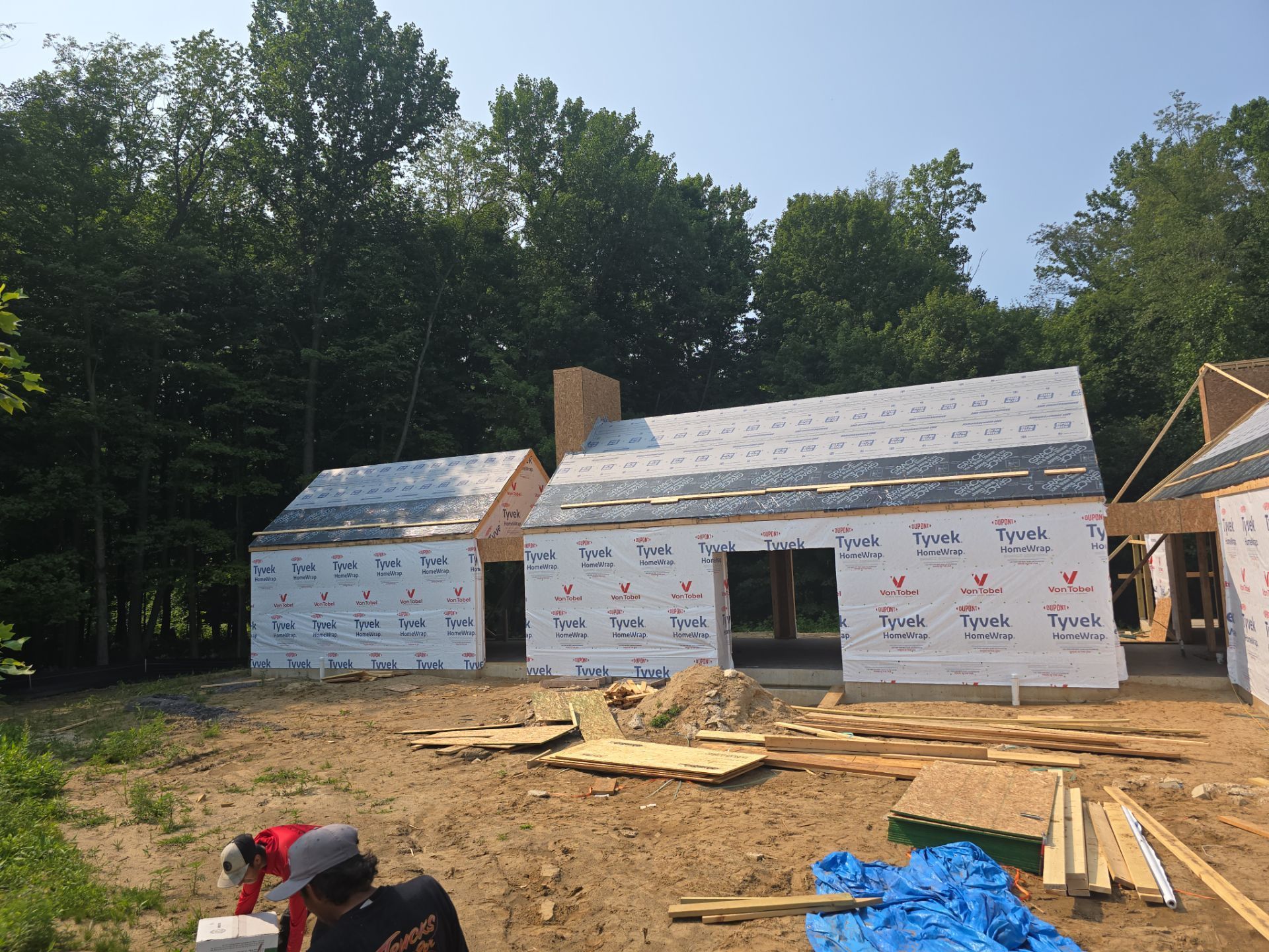 Workers constructing a house frame covered in Tyvek wrap, positioned in a wooded setting.