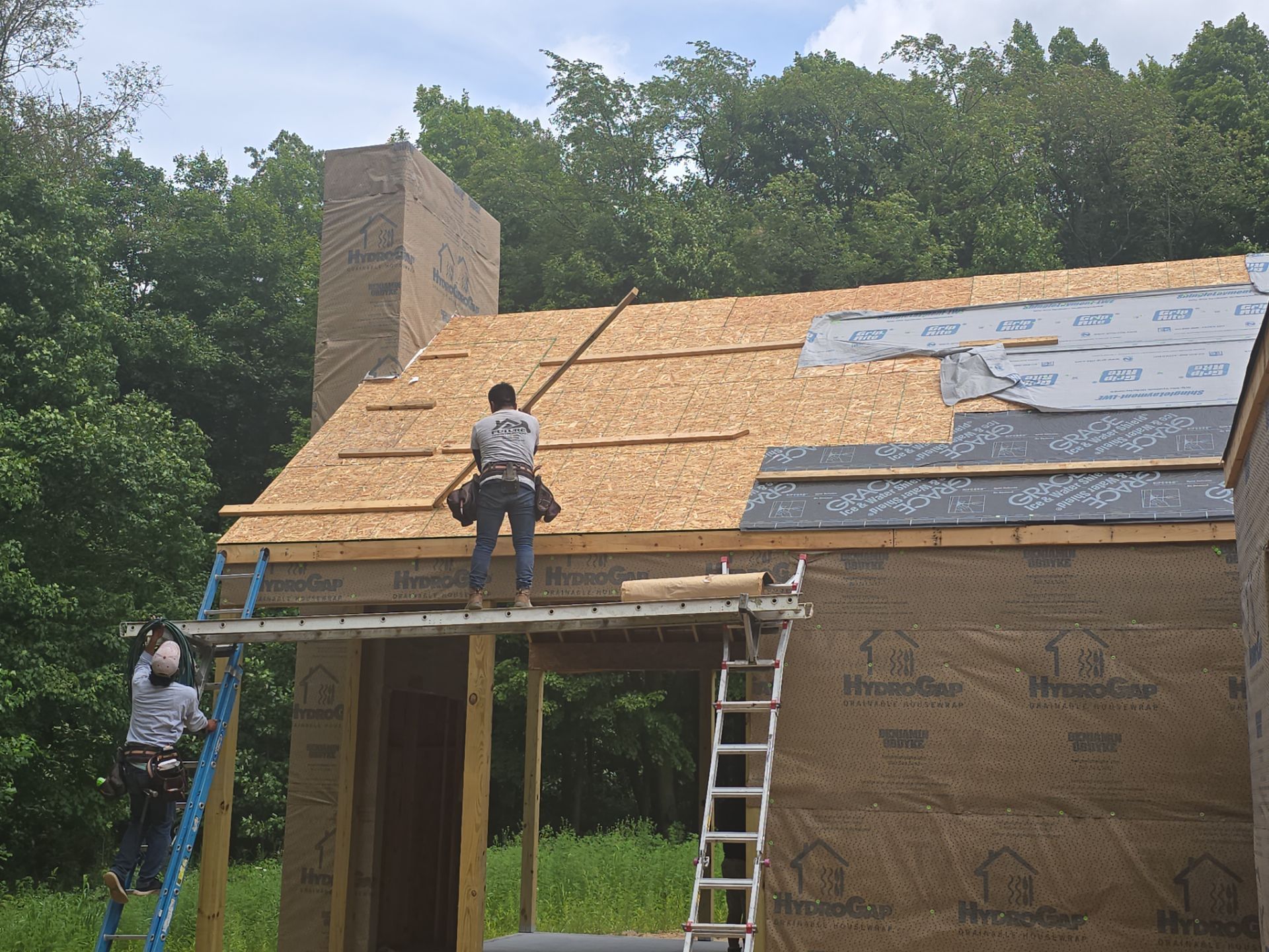 Two workers on ladders and a scaffold install plywood sheathing and roofing underlayment on a wooden house frame.