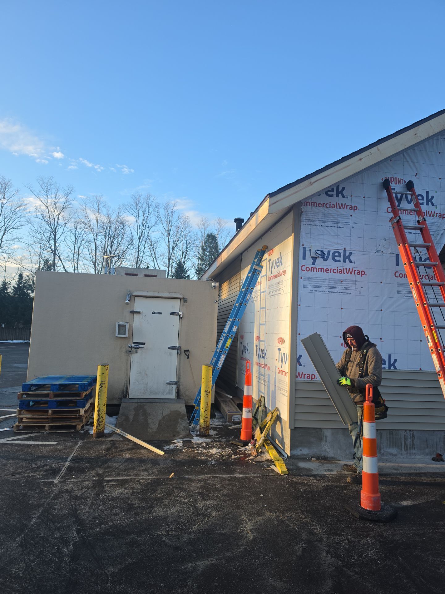 A worker installing siding on a building wrapped in Tyvek, with a ladder and outdoor freezer unit nearby.