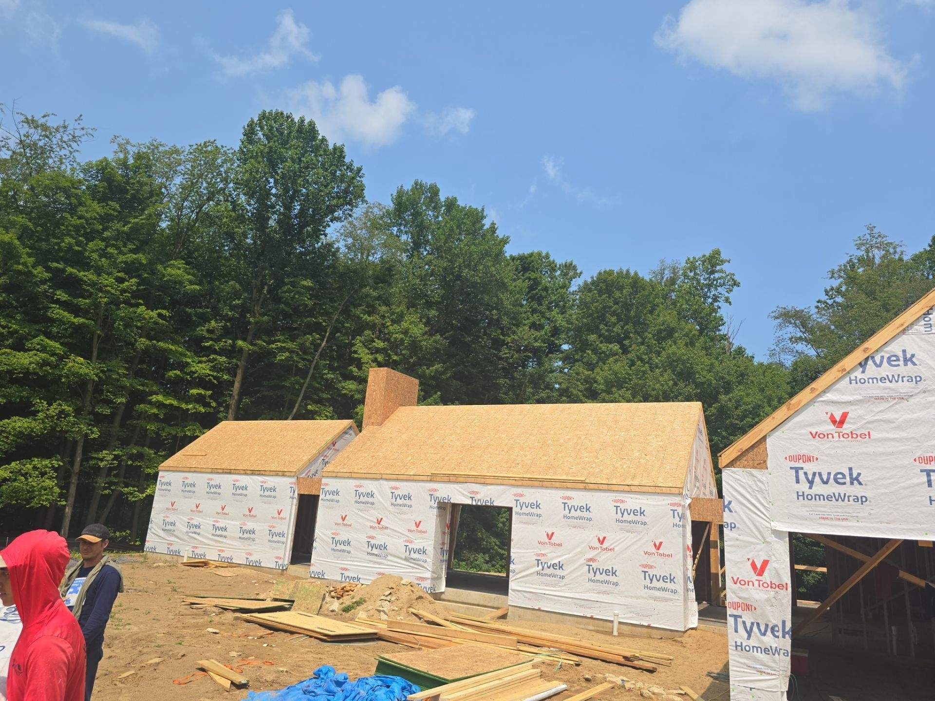 A partially constructed building frame wrapped in white Tyvek material sits in a wooded area under a blue sky.
