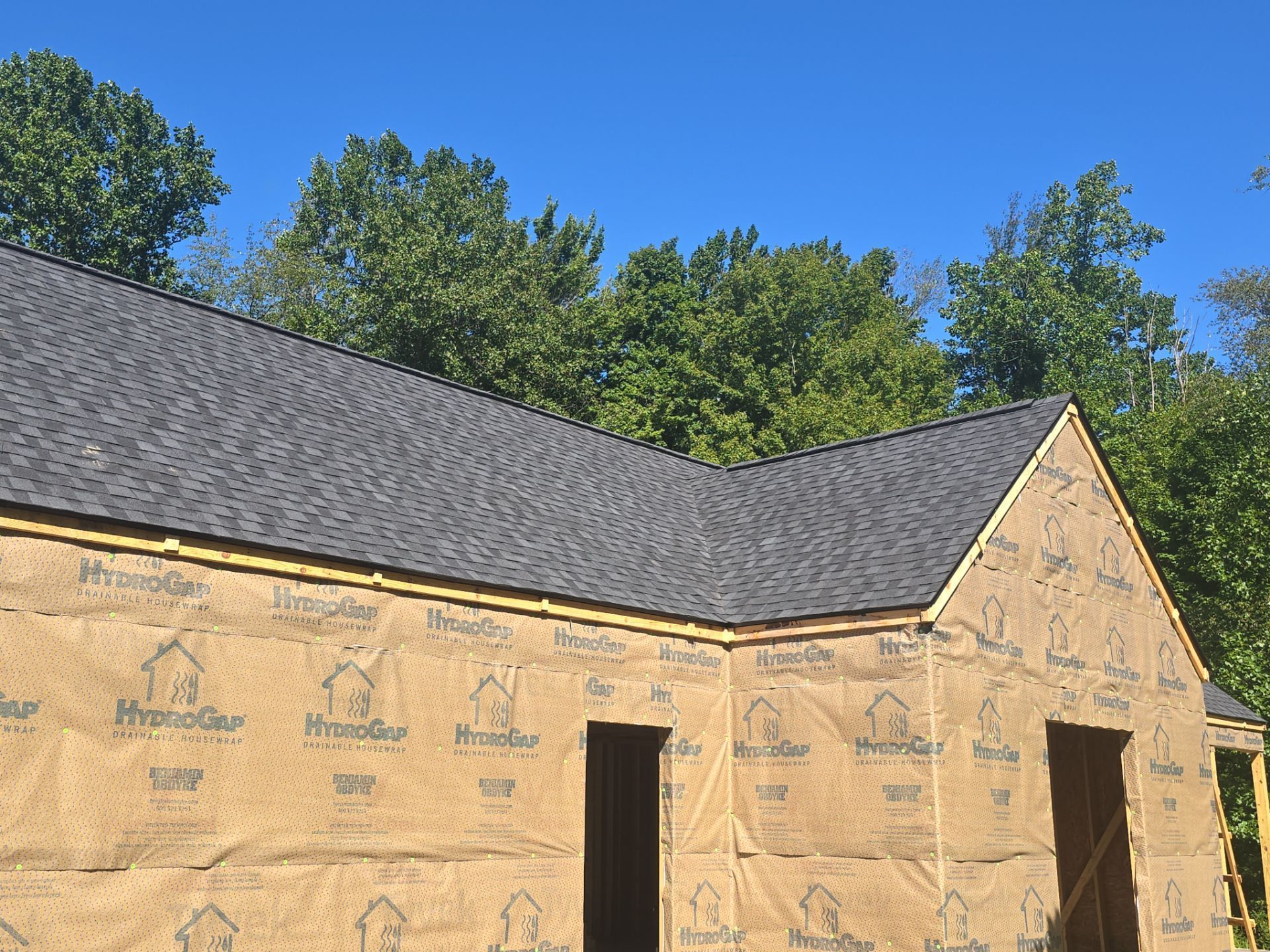A house under construction with a dark gray shingled roof and exterior walls covered in tan moisture barrier sheeting.