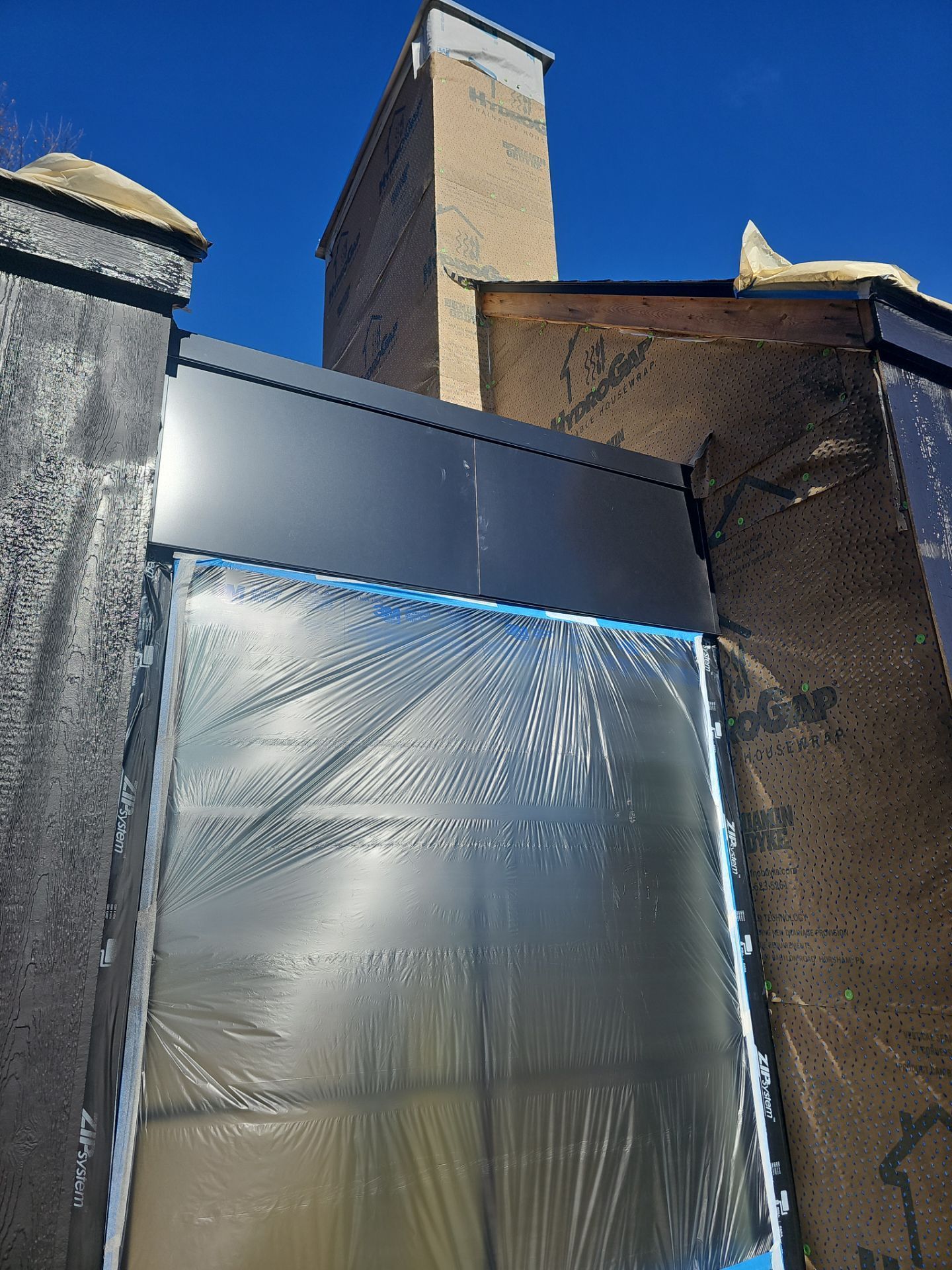 Construction site showing a window covered in protective plastic film, framed by black siding and brick wall against blue sky.