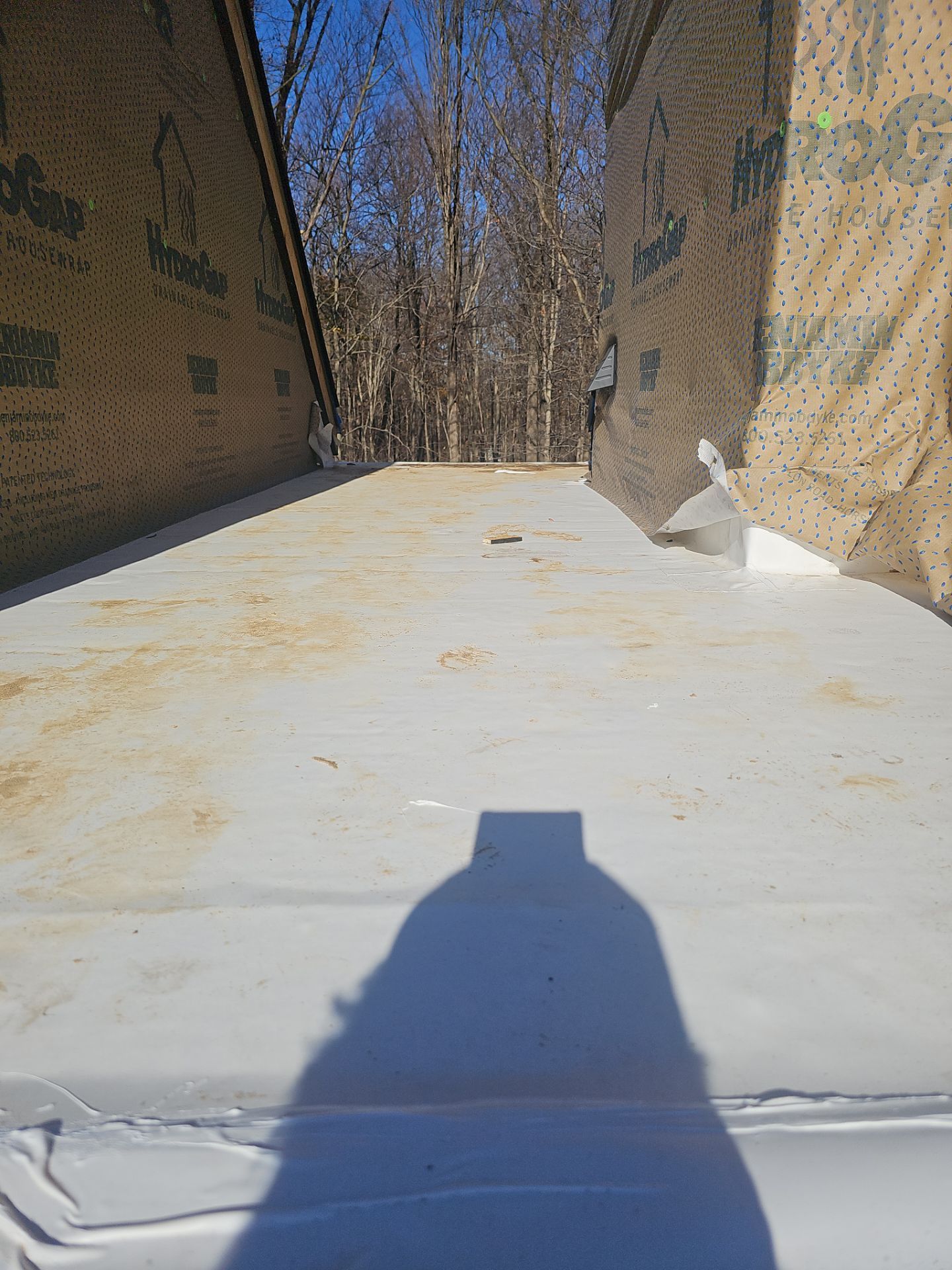 A low-slope roof with white membrane covering between two walls of building sheathing under a clear blue sky.