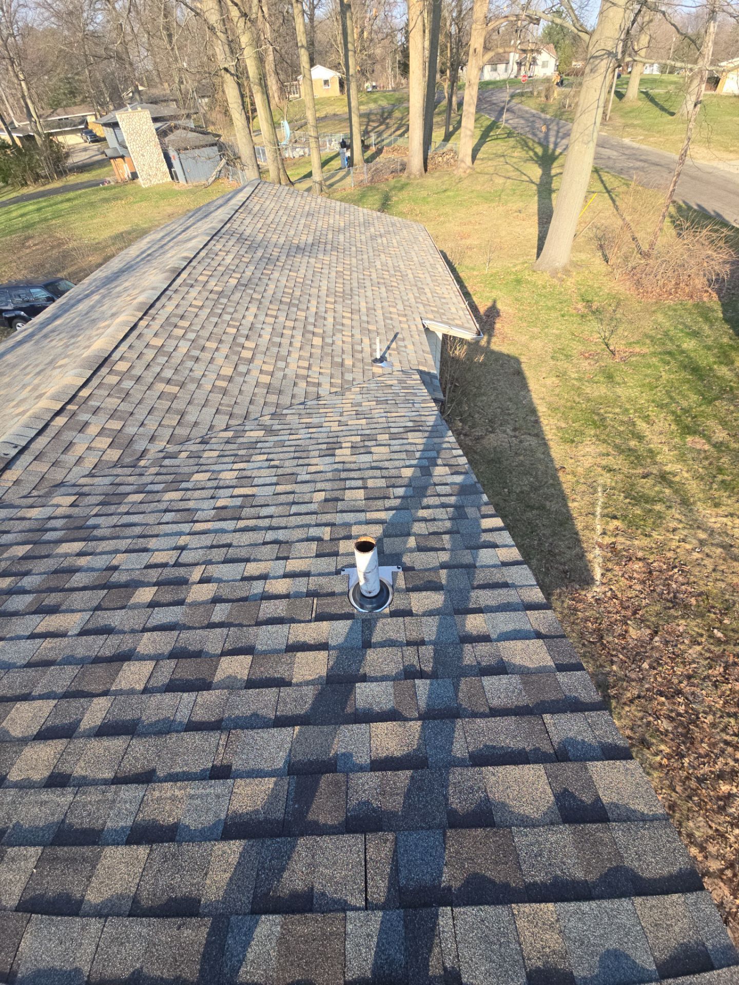 A view from a roof looking down a slope covered in grey asphalt shingles toward a wooded yard with a vent pipe visible.