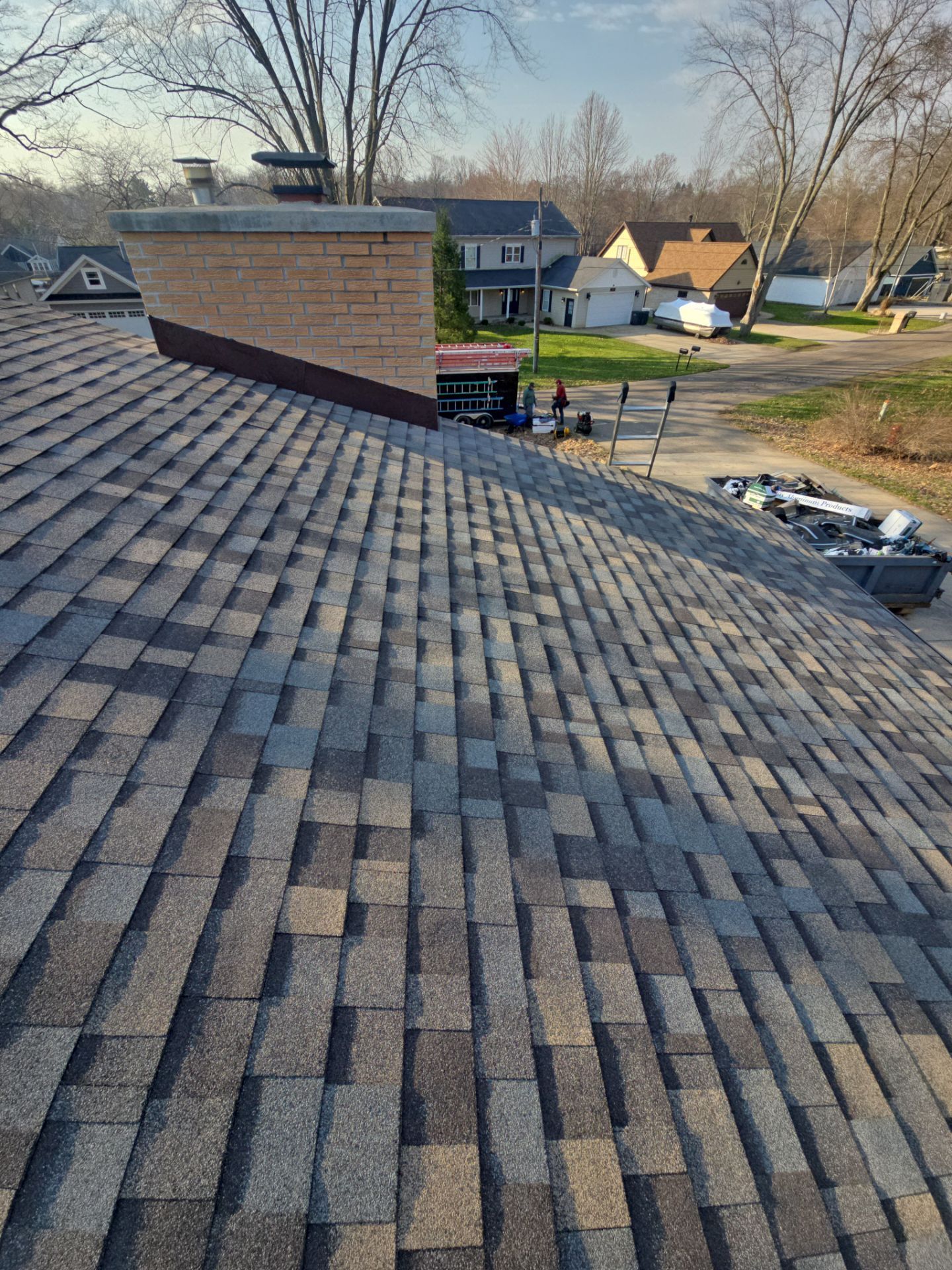 A high-angle view of a newly shingled gray asphalt roof, featuring a brick chimney with a metal cap and a dumpster below.