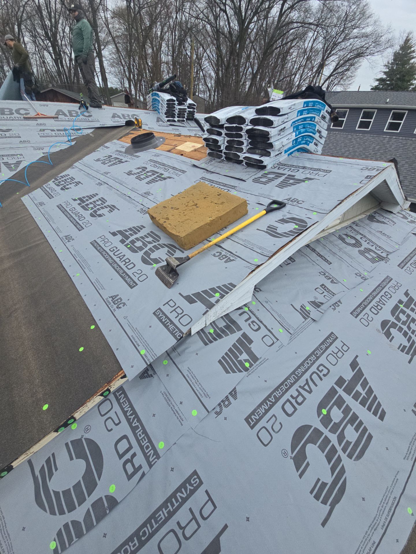 Workers install synthetic underlayment on a residential roof with a stack of shingles and tools nearby.
