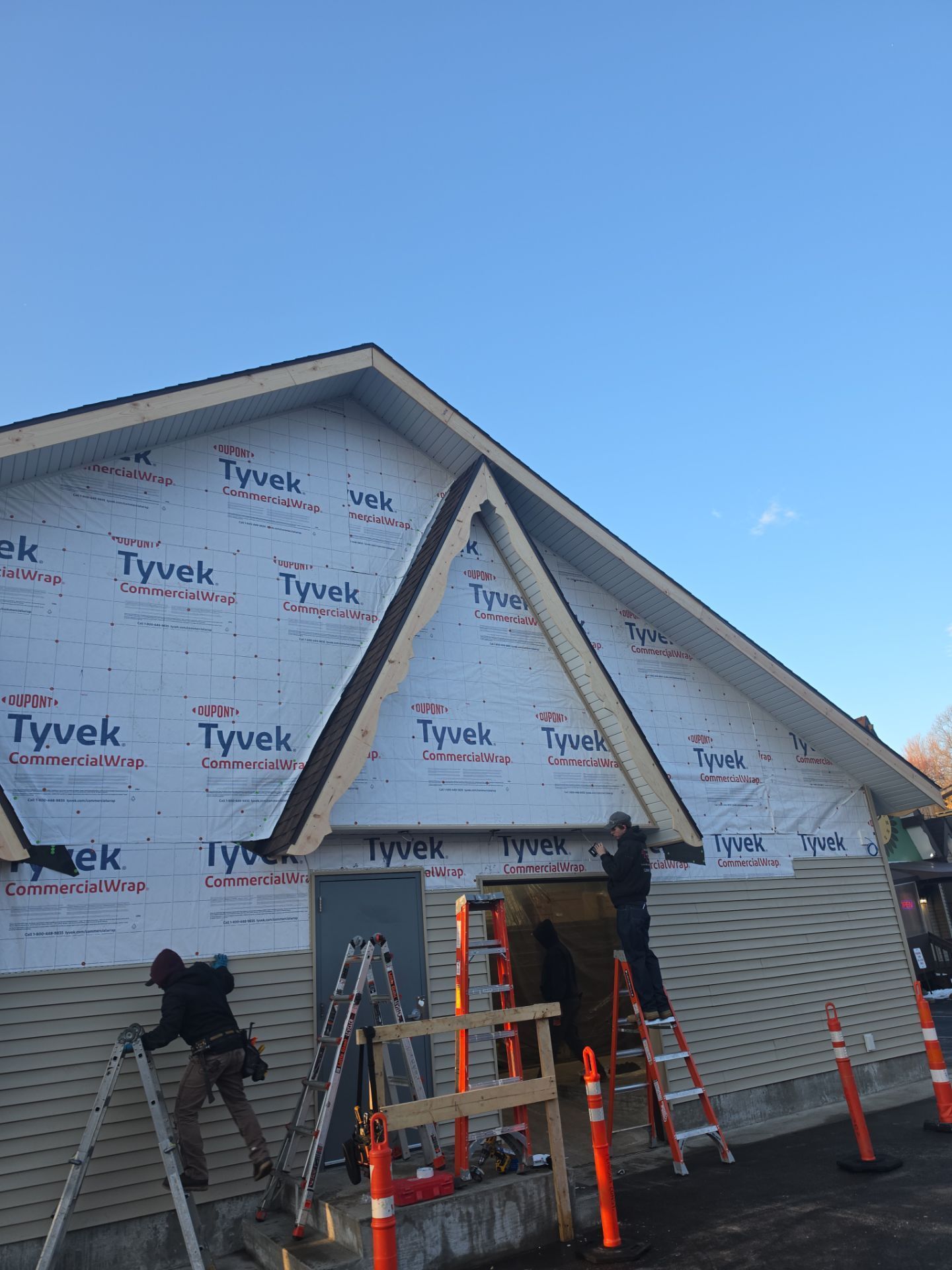 Two workers install siding on the exterior of a building covered in Tyvek wrap, working on ladders under a bright blue sky.