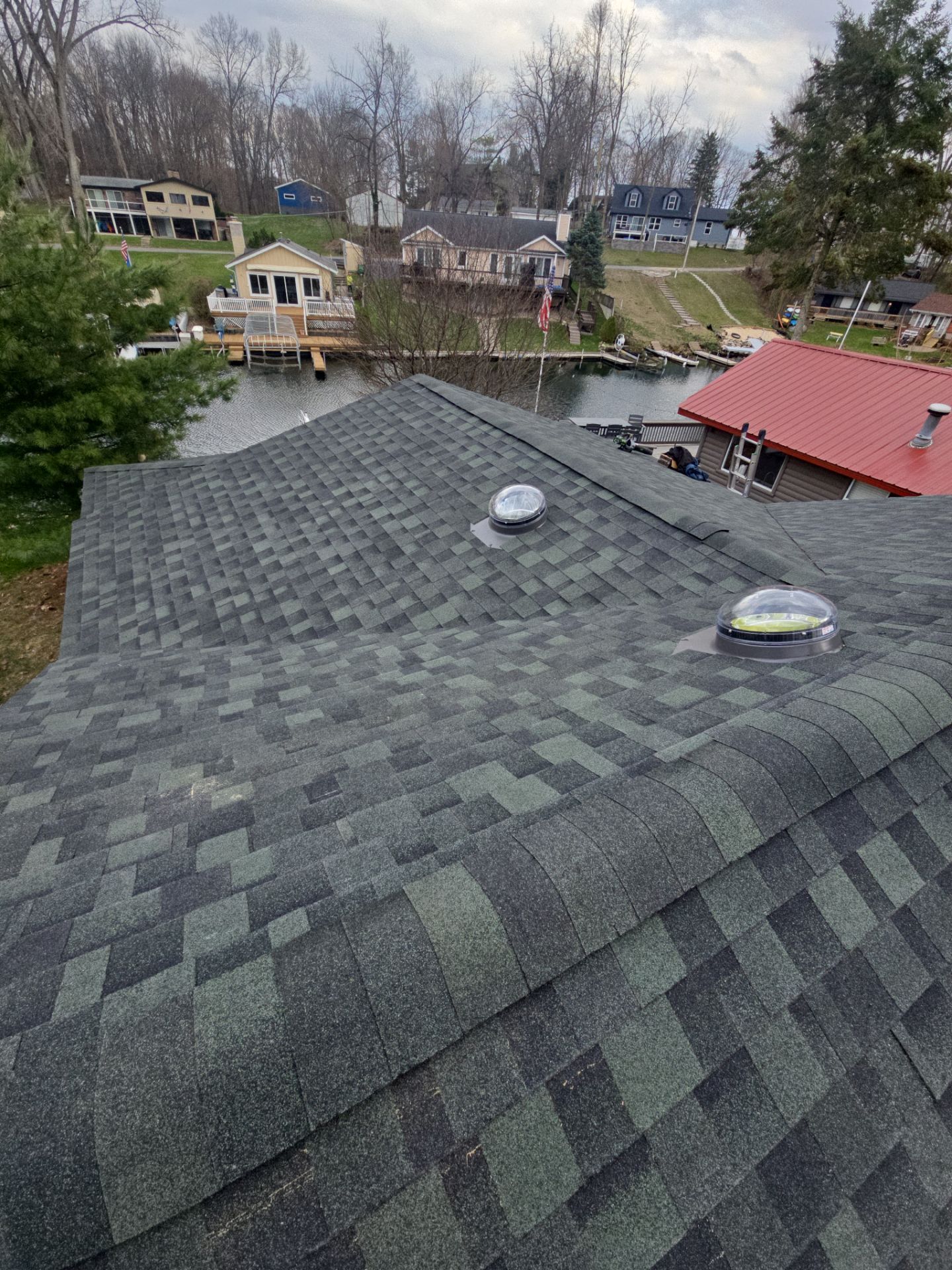 A view from above looking over a dark-shingled roof with two skylights toward a lake with houses on the opposite shore.