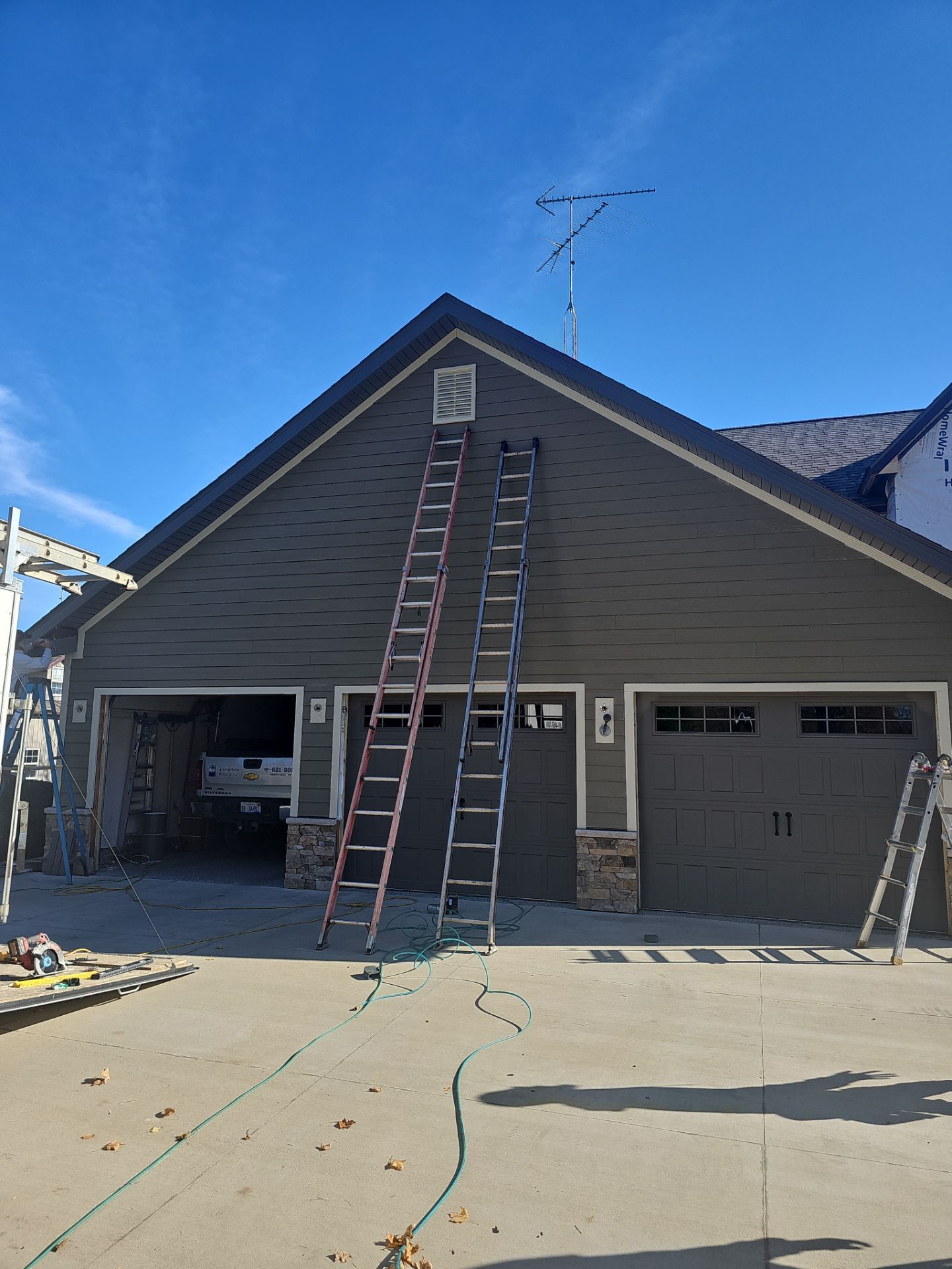 Two tall ladders lean against the dark gray siding of a three-car garage under a bright blue sky.