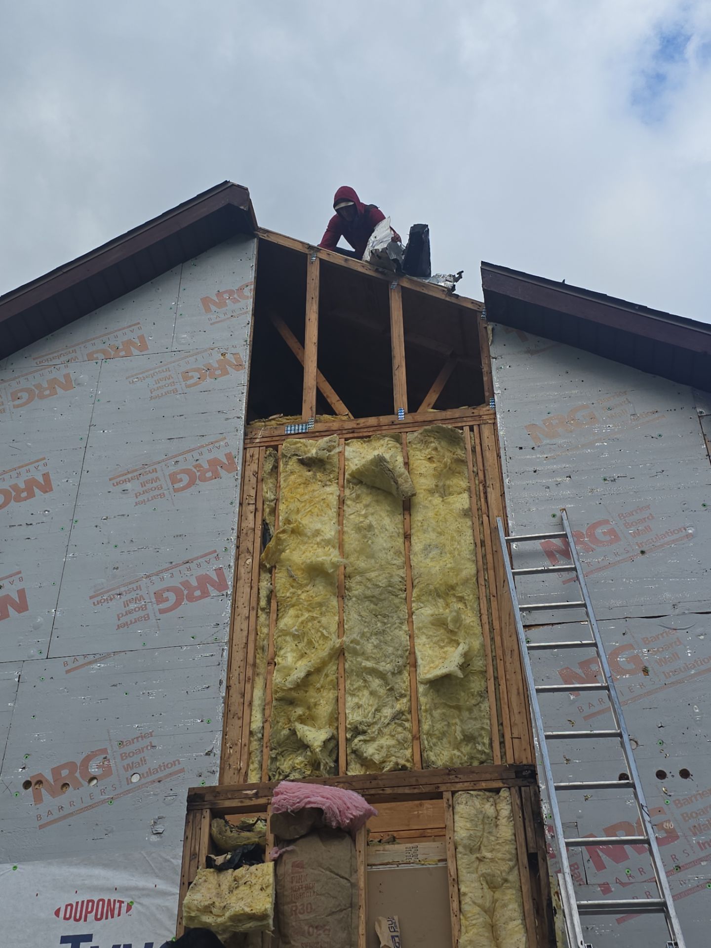 A worker stands atop a framed exterior wall section of a house, which has exposed insulation and a nearby leaning ladder.