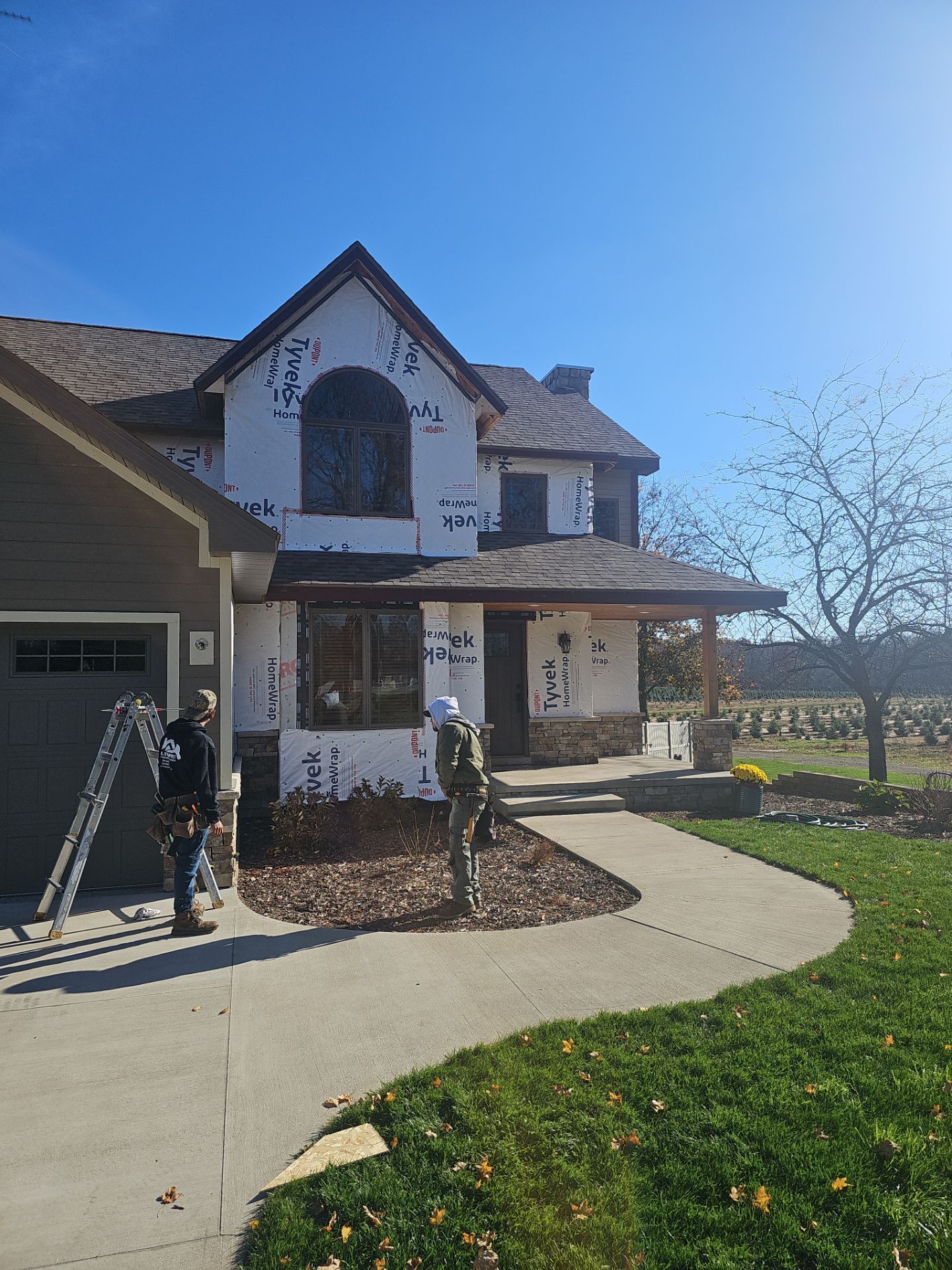 Two workers in work gear stand in front of a house under construction with exposed sheathing and an aluminum ladder.