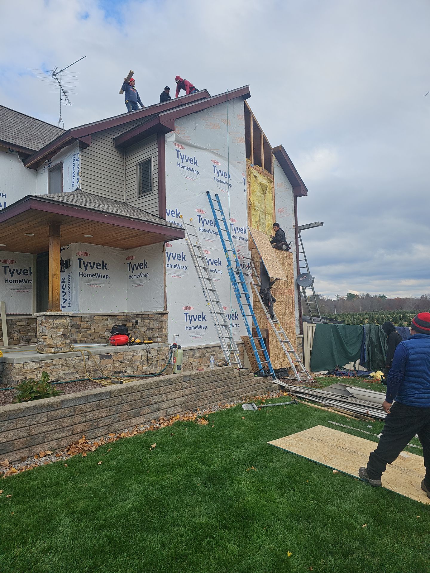 Construction workers on a roof and ladders renovating the exterior of a multi-story house with Tyvek wrapping.