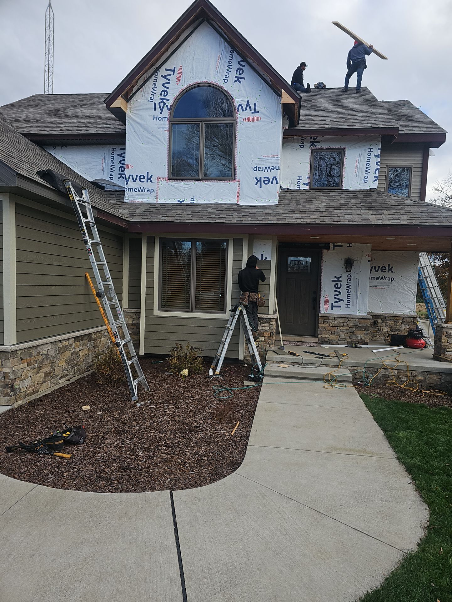Contractors in work gear repair the roof and siding of a two-story home partially wrapped in Tyvek protective sheeting.