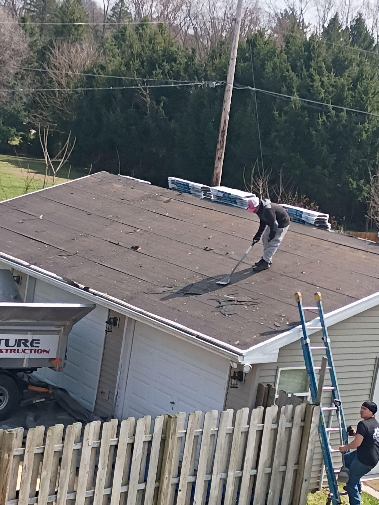 A worker stands on a garage roof stripping shingles, while another worker stands on a ladder nearby.