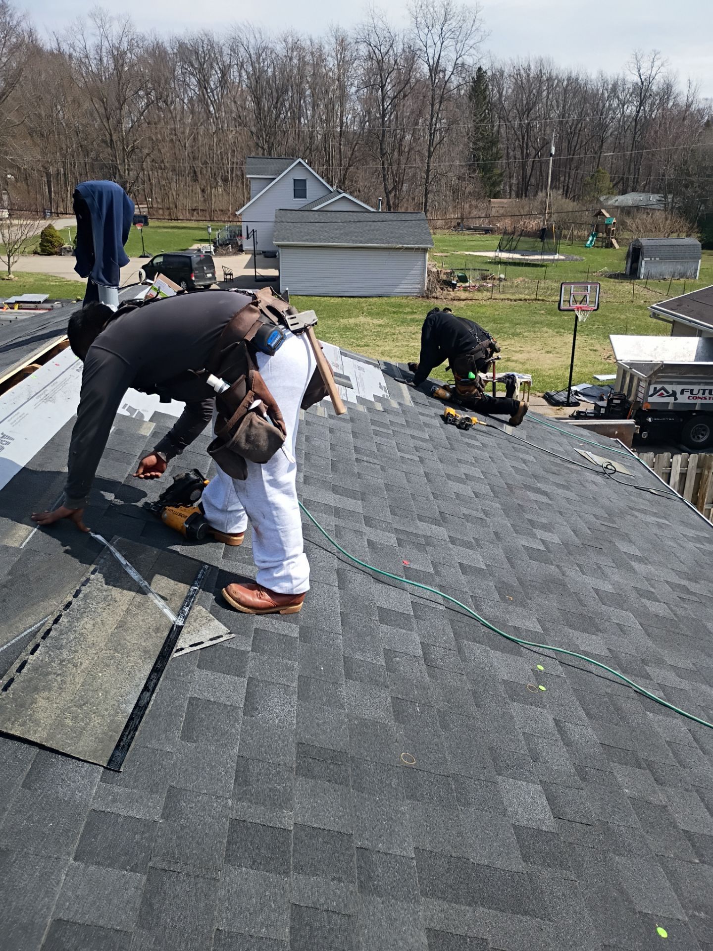 Two people in work clothes install dark shingles on a sloped residential roof under a clear sky.