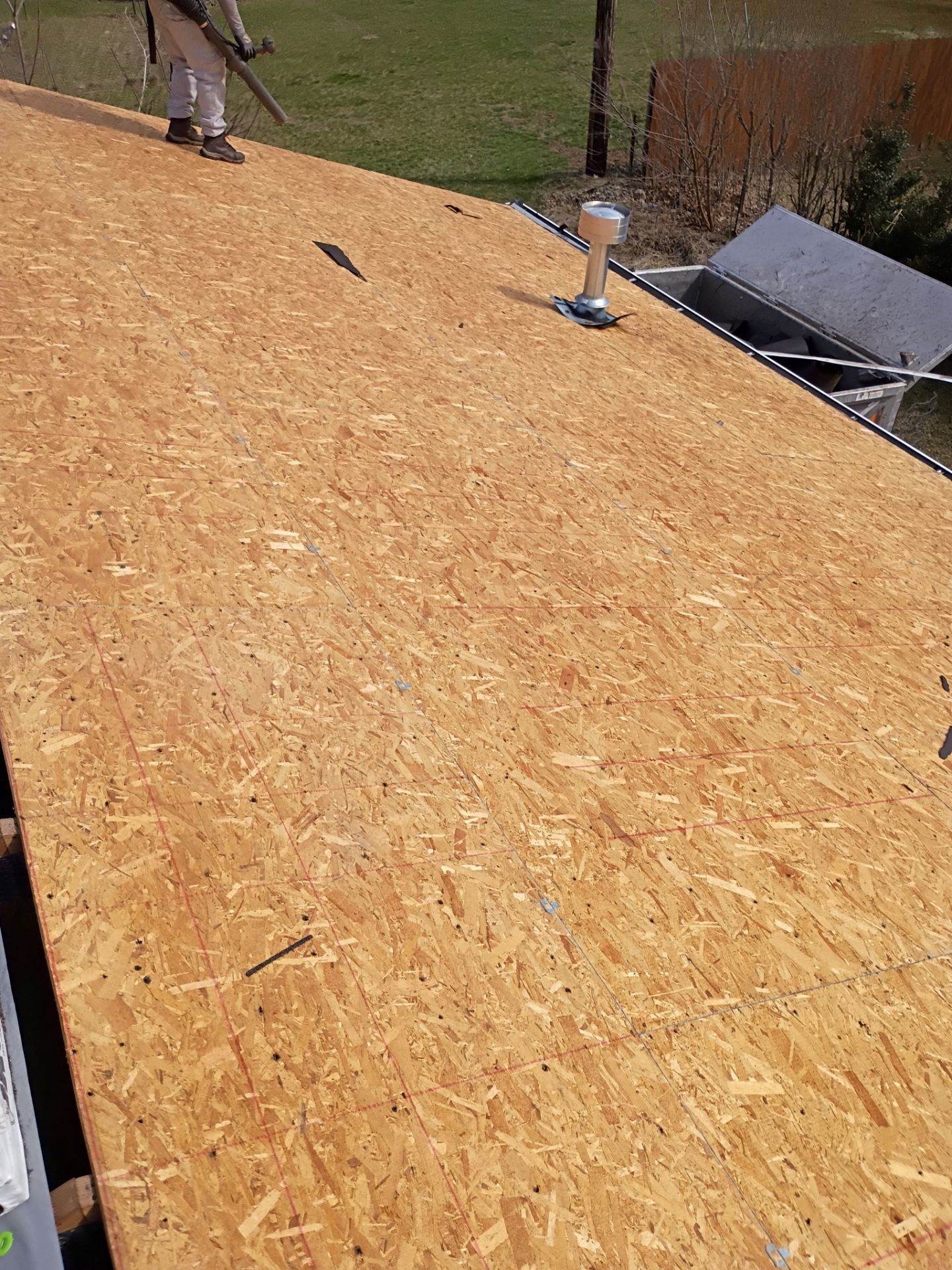 A person with a leaf blower cleans an unfinished wooden roof deck with a plumbing vent pipe extending from the surface.