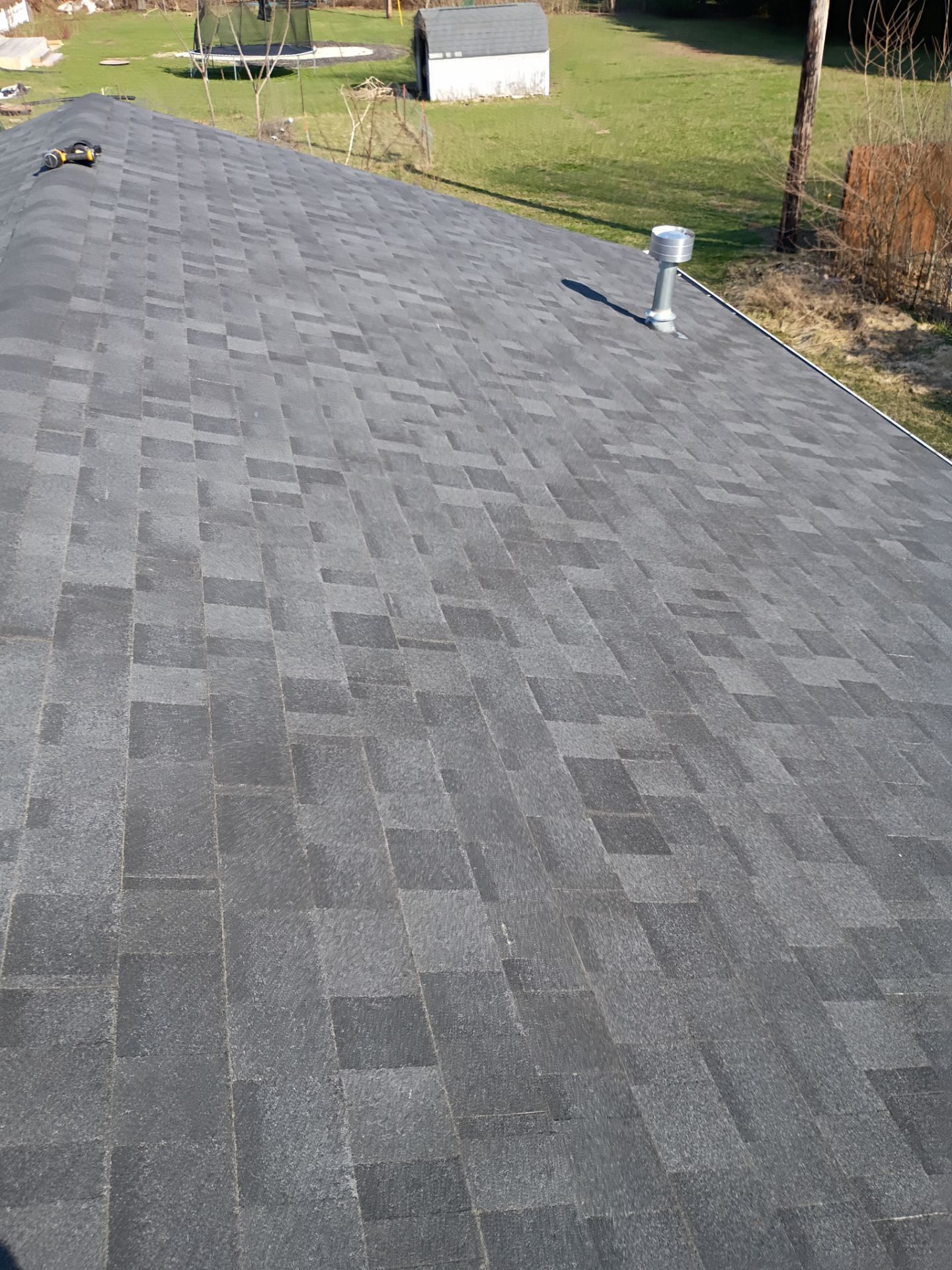 An elevated view of a shingled roof with a metal ventilation pipe, overlooking a grassy yard with a small shed.