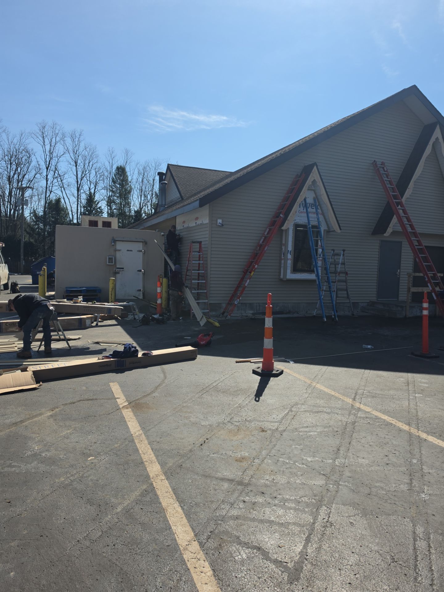 Construction workers renovate the exterior of a building with ladders, scaffolding, and traffic cones in a parking lot.