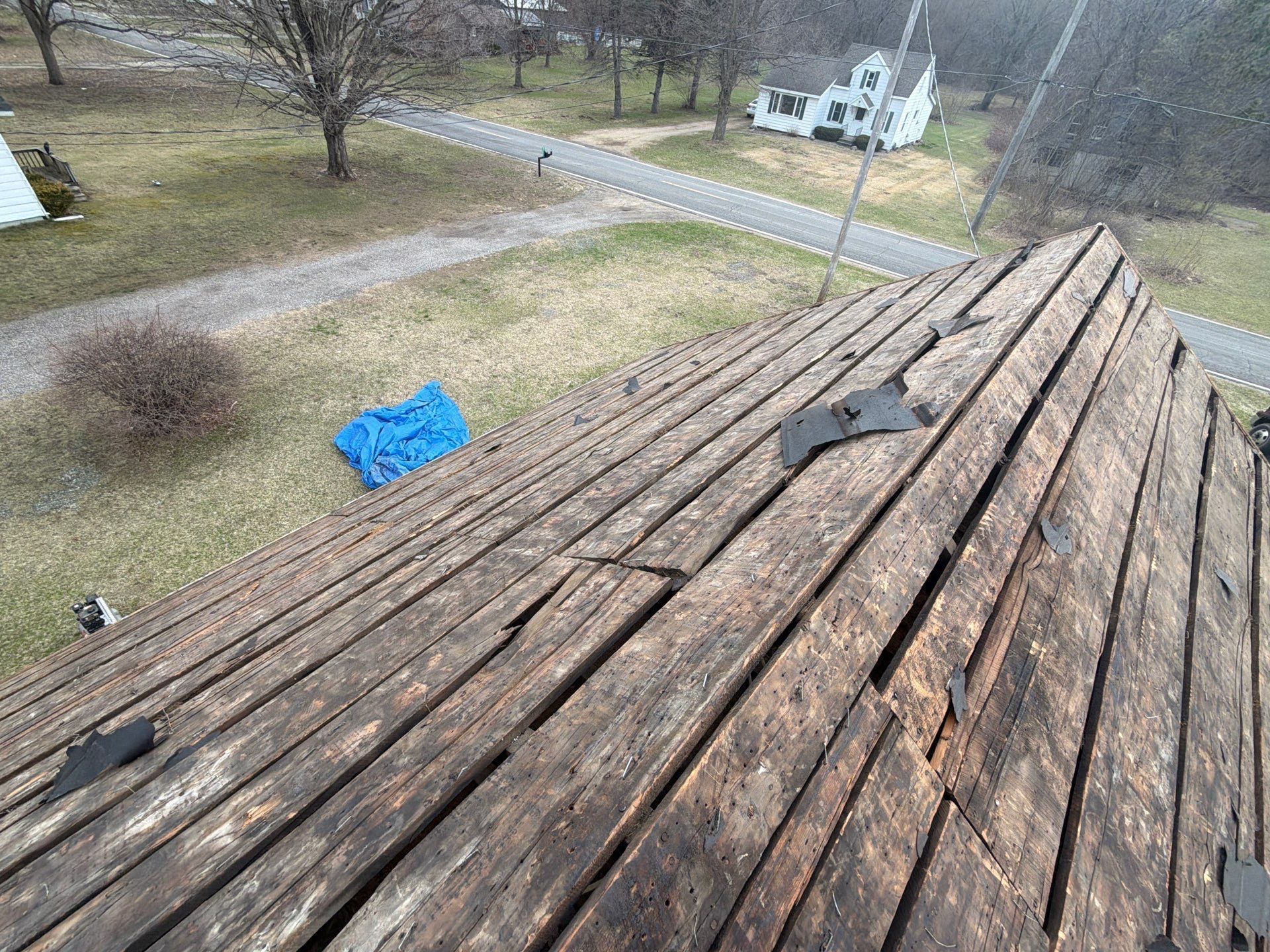 A high-angle view of a wooden roof deck with stripped shingles, showing planks under a clear sky and distant house.
