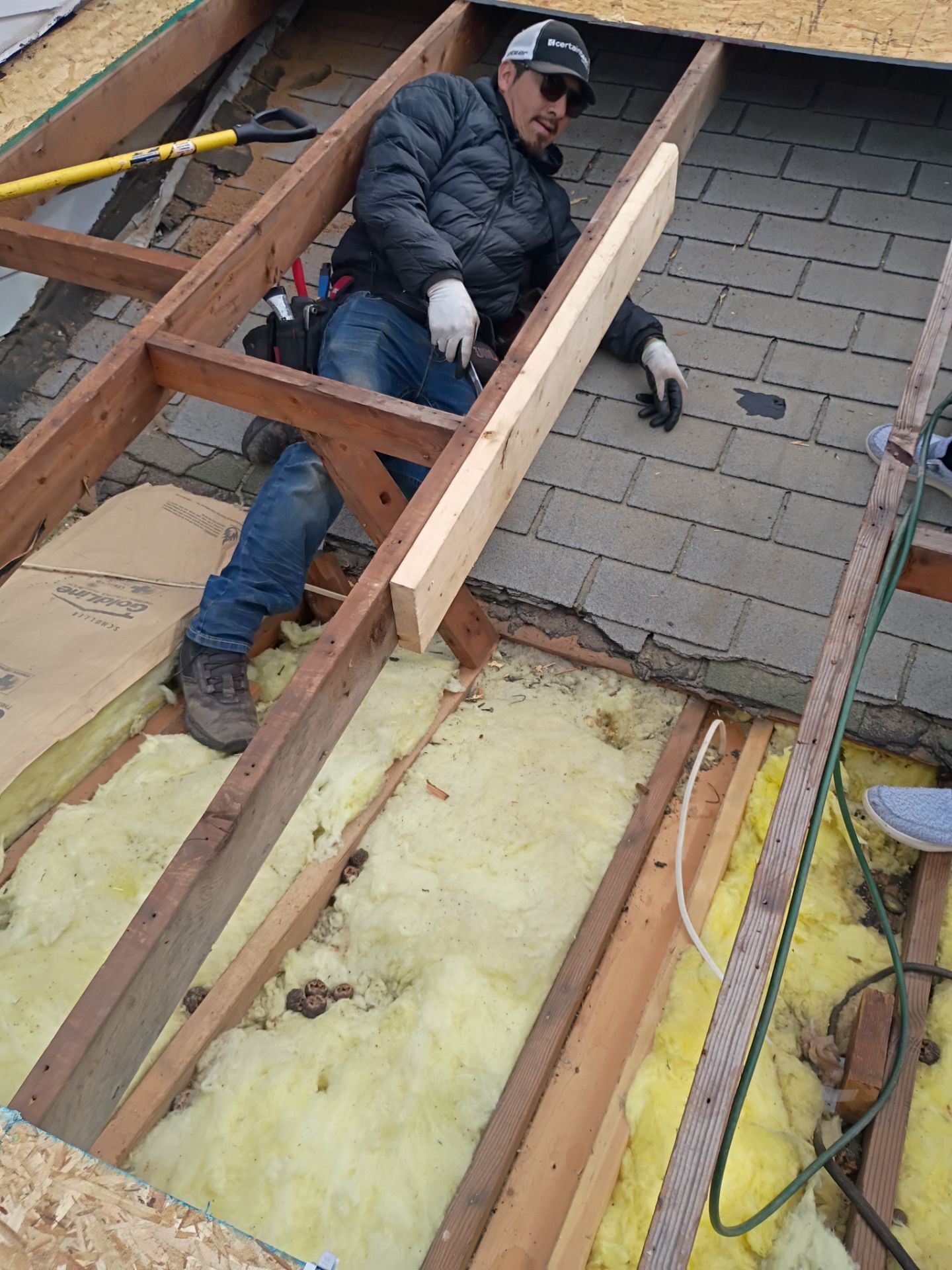 A worker wearing a cap and jacket sits in a rooftop opening, installing wooden framing above yellow fiberglass insulation.