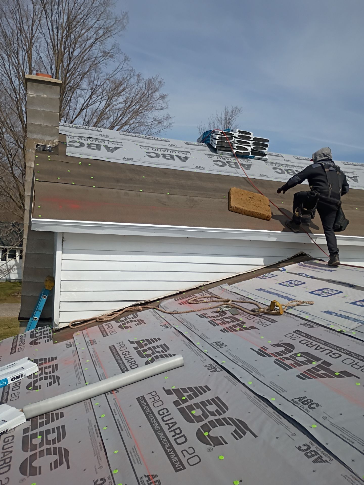 A person wearing a harness performs roofing work on a house covered in ABC-branded underlayment under a blue sky.