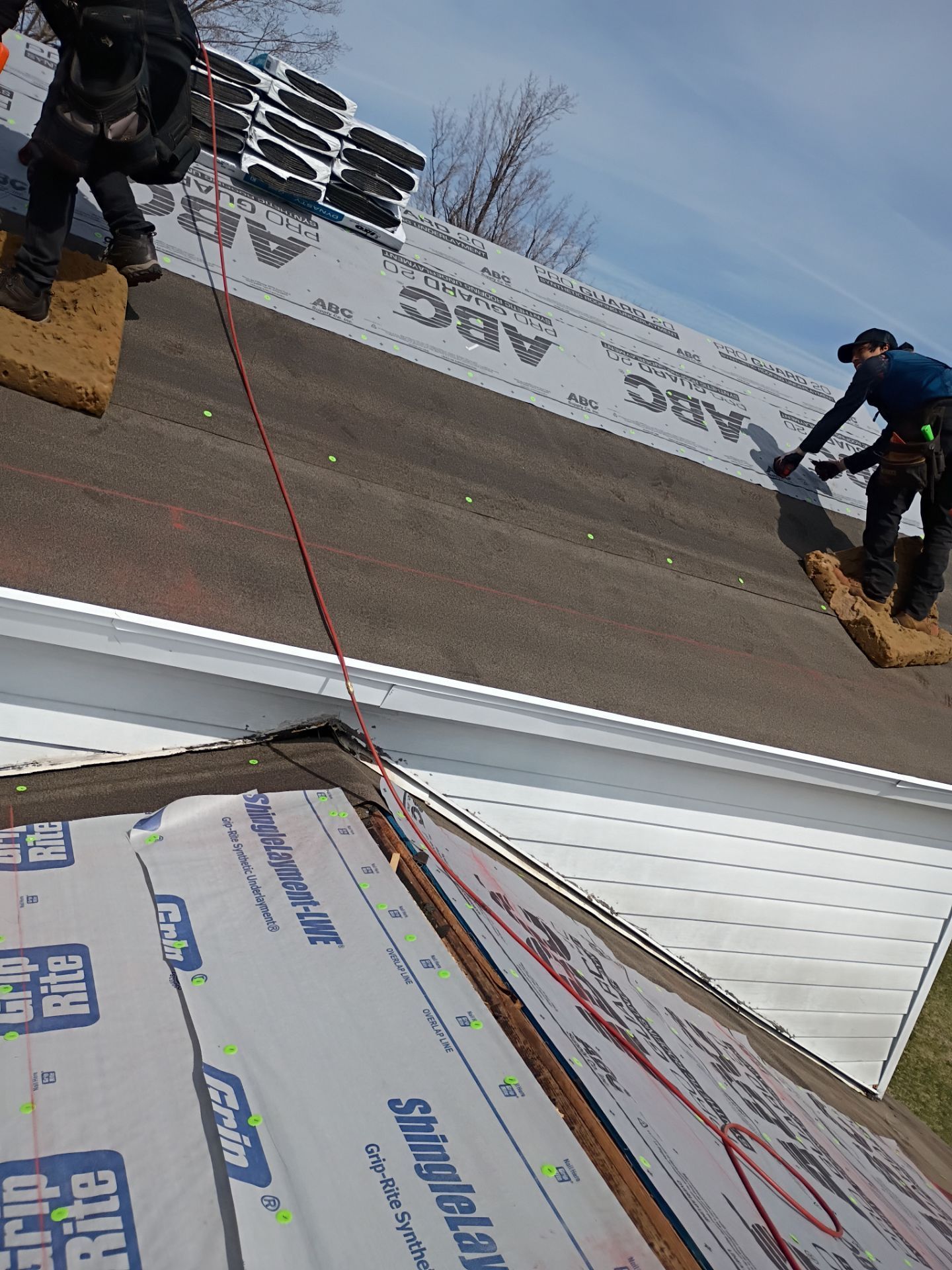 Two roofers stand on a sloped roof underlayment, working on a residential roofing project on a sunny day.