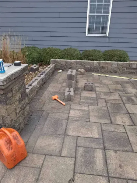 Patio construction: Grey pavers with stacked blocks and a mallet, beside a blue house and short wall.