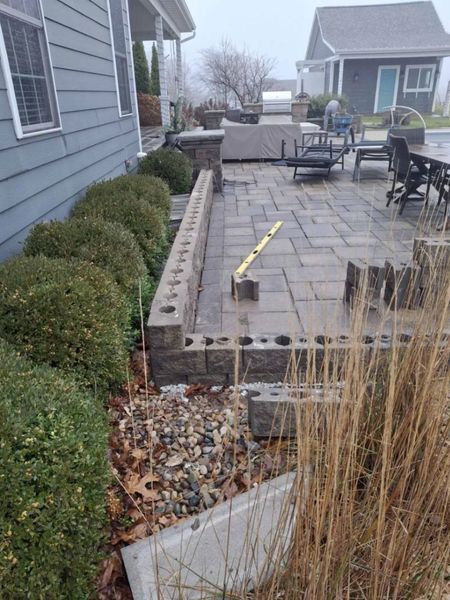 A partially built patio with a retaining wall, bushes, and gray pavers next to a blue house.
