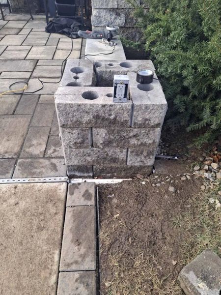 Stone block structure being built on patio with pavers, surrounded by dirt and shrubbery.