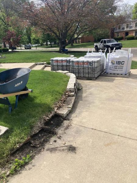 A wheelbarrow, paving materials, and bags sit beside a driveway, suggesting a landscaping project is underway.