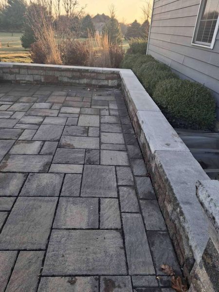 Stone patio with low brick wall, next to a house and landscaping.