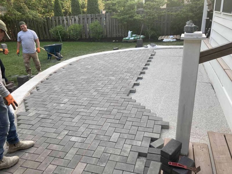 Workers laying gray brick pavers for a patio; grass and fence in the background.