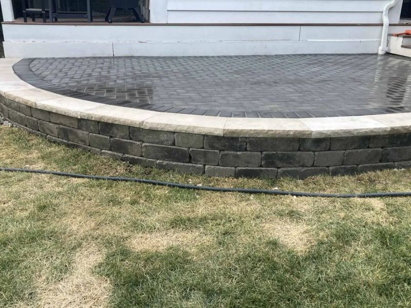 Curved brick patio with retaining wall, grass in foreground, house in background.