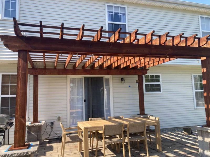 Wooden pergola over patio with a table and chairs set. Back of a light-colored house in the background.