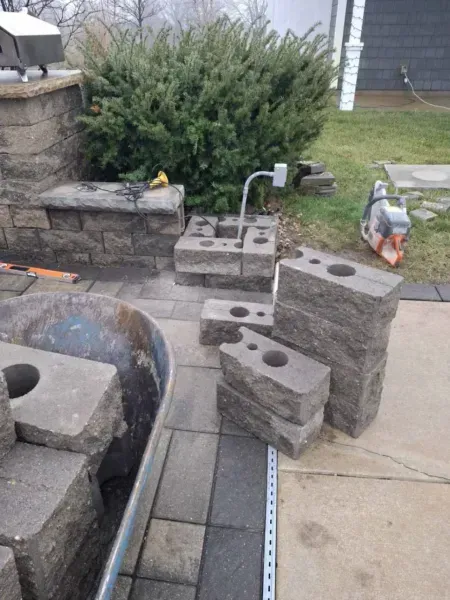 Building a patio with retaining wall blocks. Concrete blocks stacked on a gray patio, next to a bush.