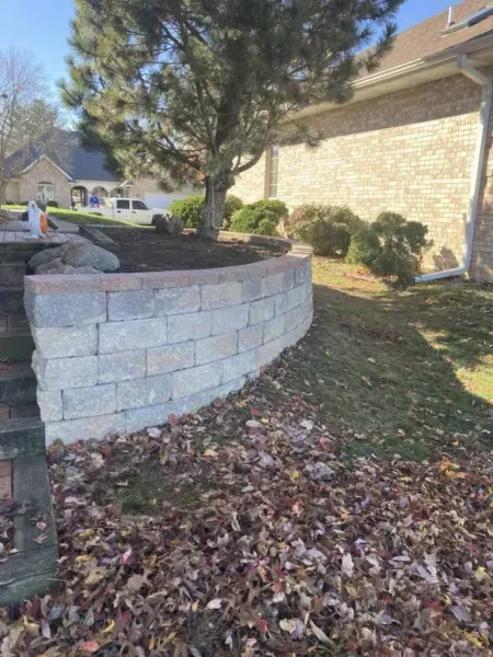 A curved retaining wall made of gray blocks supports a tree and landscaping near a brick building.