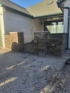 Stone pillars frame a gravel patio, leading to a house with a dark door.