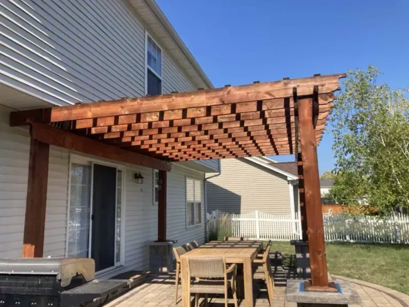 Wooden pergola over a patio with a dining table and chairs, attached to a two-story house.