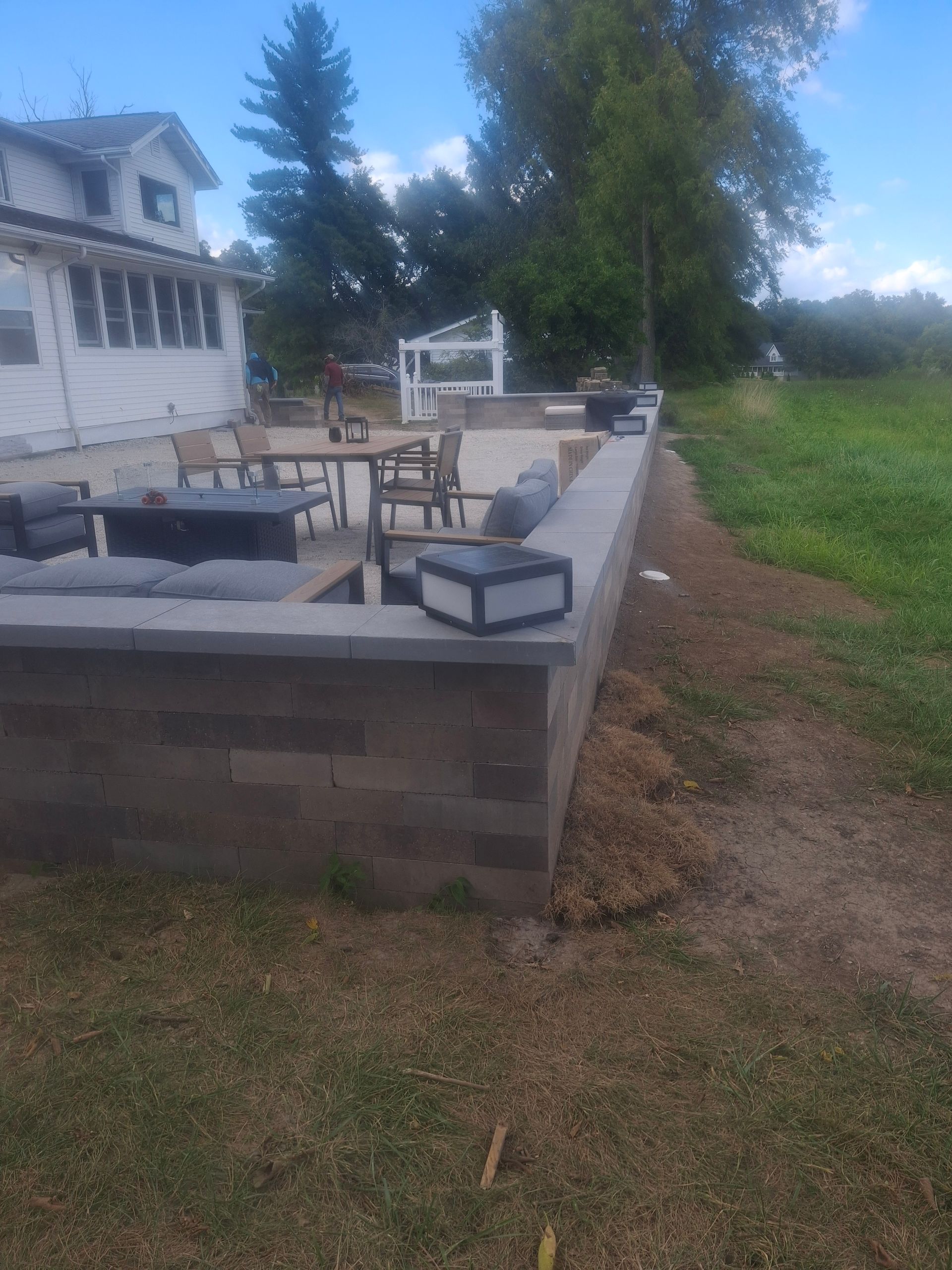 A stone patio with outdoor furniture, a fire pit, and solar lights, next to a grassy area and a house.
